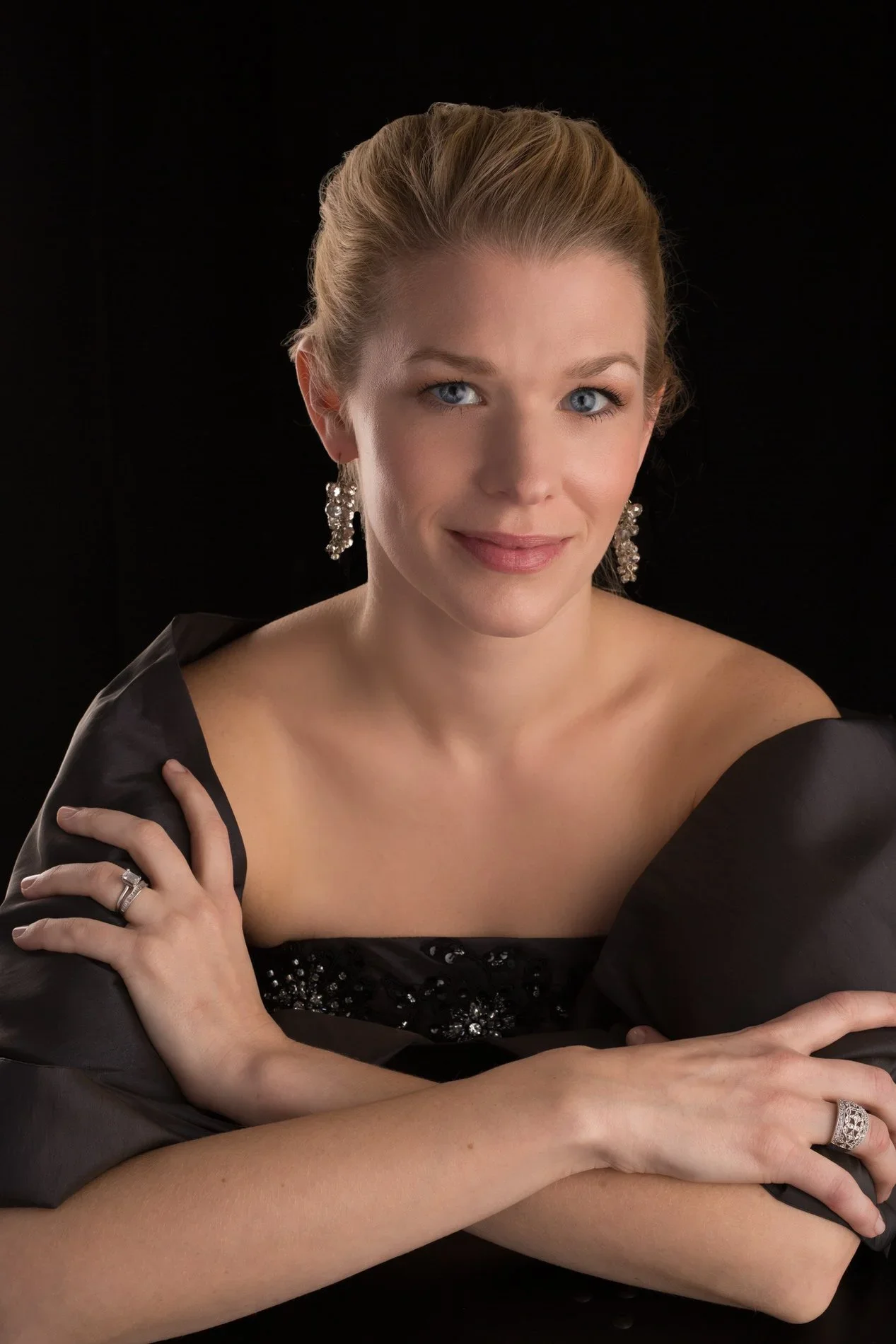 Portrait of a woman with blonde hair styled up, blue eyes, wearing earrings and a black gown with textured embellishments, against a black background.