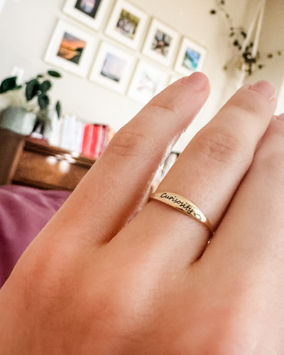 A hand wearing a ring with the word 'curiosity' engraved on it, set against a blurred background of a home interior with framed pictures on the wall.