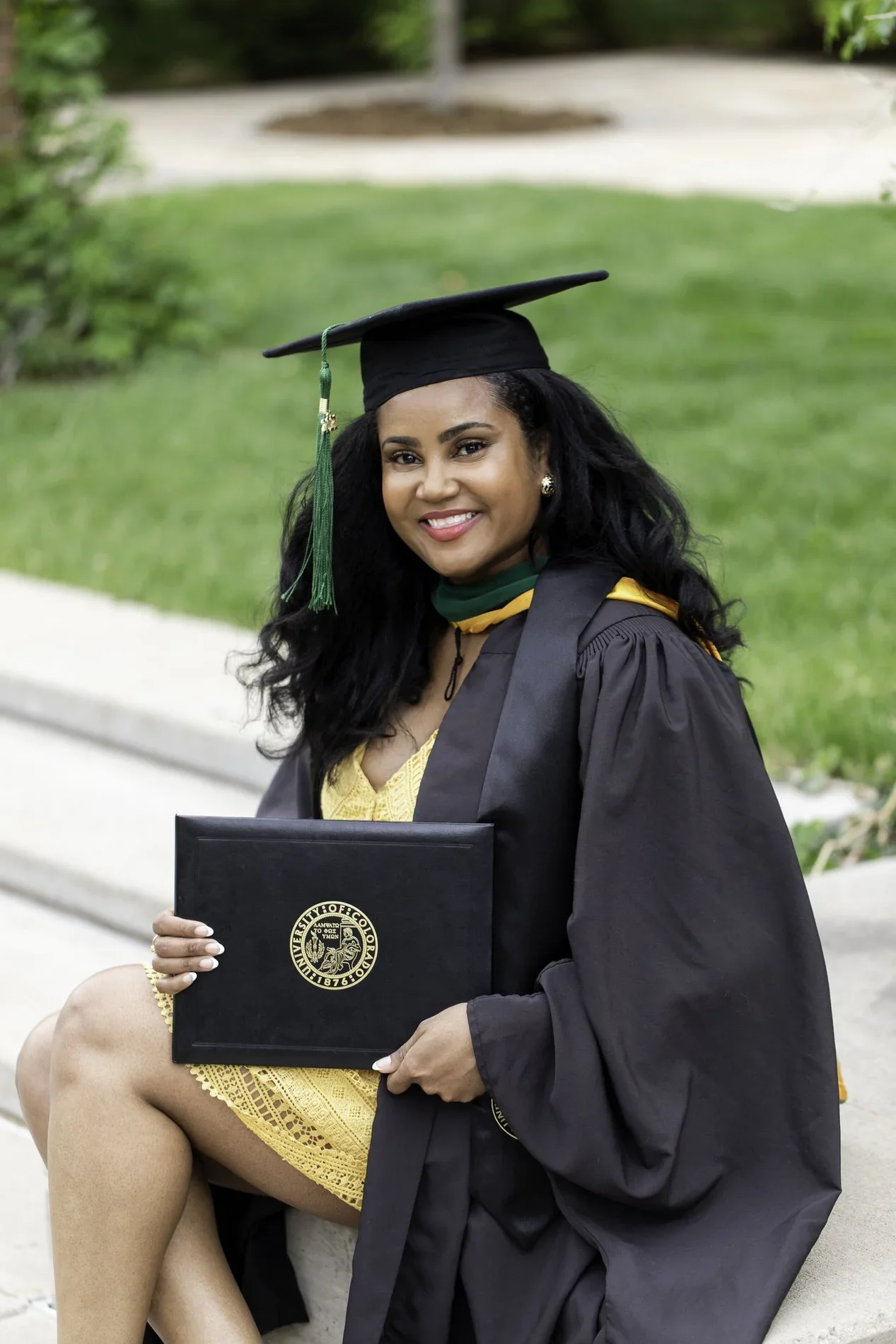 A woman in a graduation cap and gown sitting outdoors, holding a diploma, smiling, with greenery and sidewalk in the background.