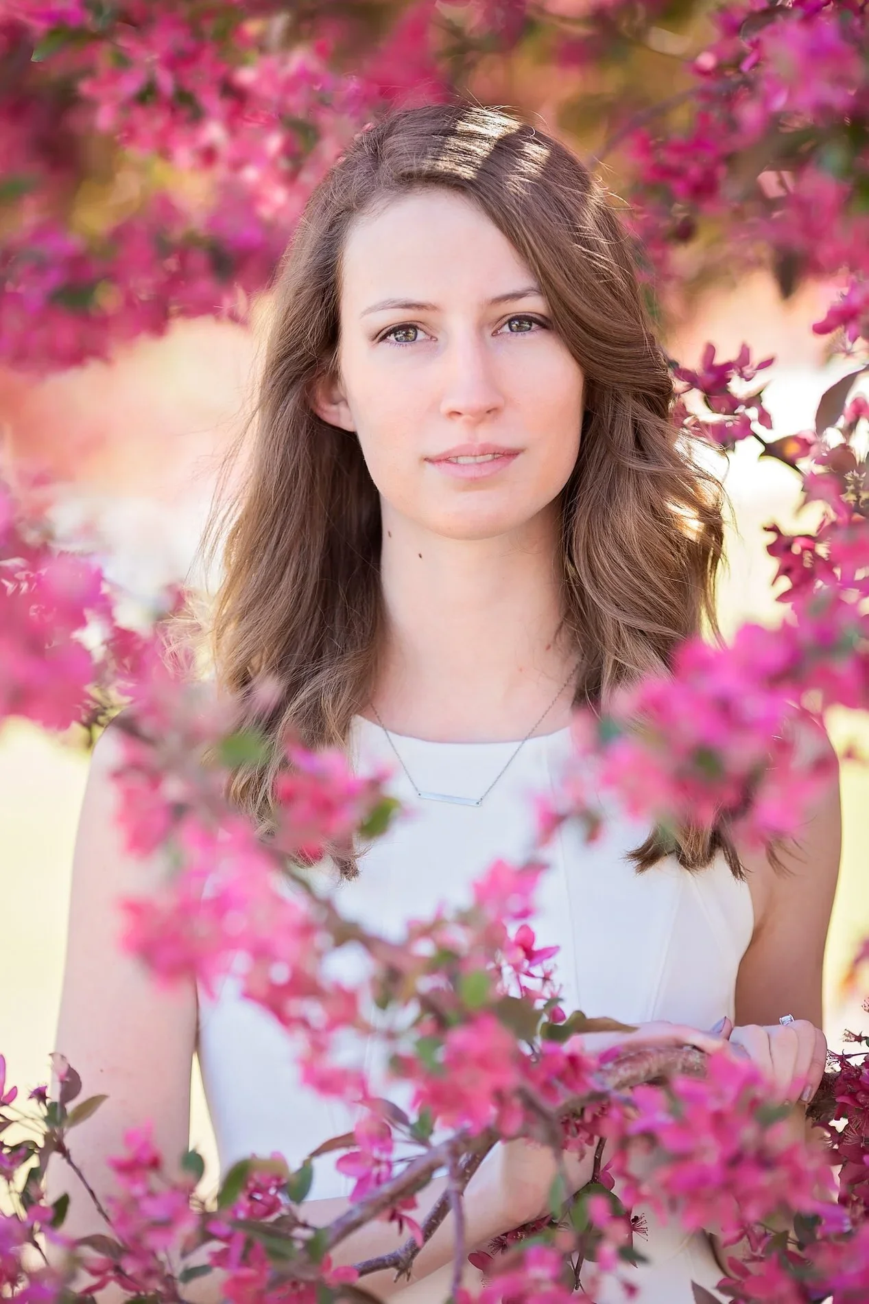 A young woman with long wavy brown hair and light skin, standing among pink flowering branches, wearing a white sleeveless top and a delicate necklace outdoors during daytime.