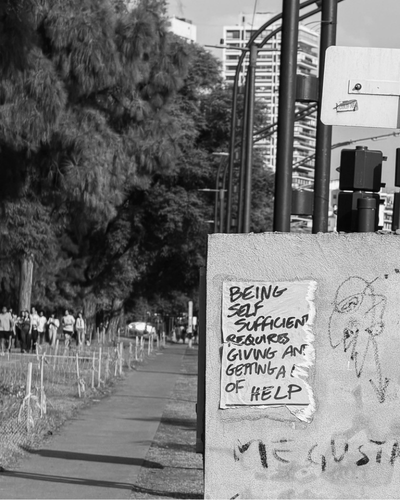Sight of a sidewalk with a group of people walking, large trees lining the street, and a concrete utility box with graffiti and a handwritten note about self-sufficiency and asking for help.