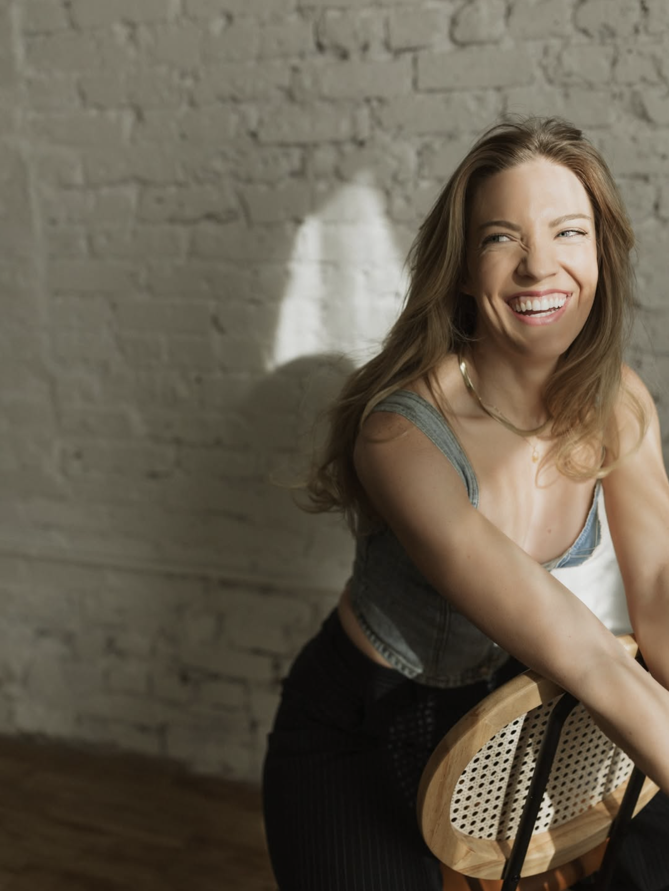 A young woman with long brown hair, smiling and looking to her left, sitting in front of a brick wall. She is wearing a sleeveless top and a necklace. A wooden chair with a woven backrest is in front of her.
