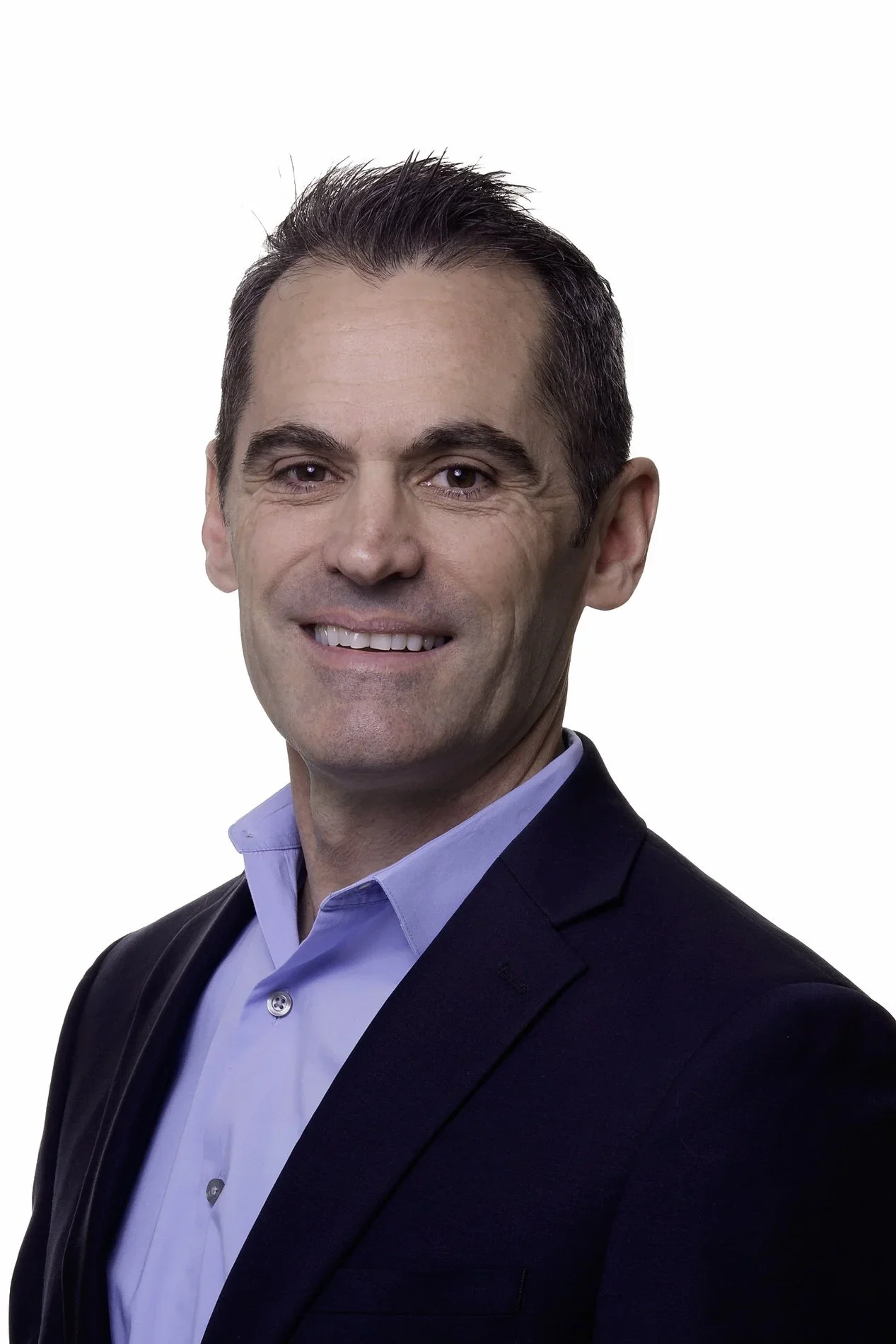 Headshot of a smiling man with short dark hair, wearing a blue collared shirt and a black blazer, against a white background.