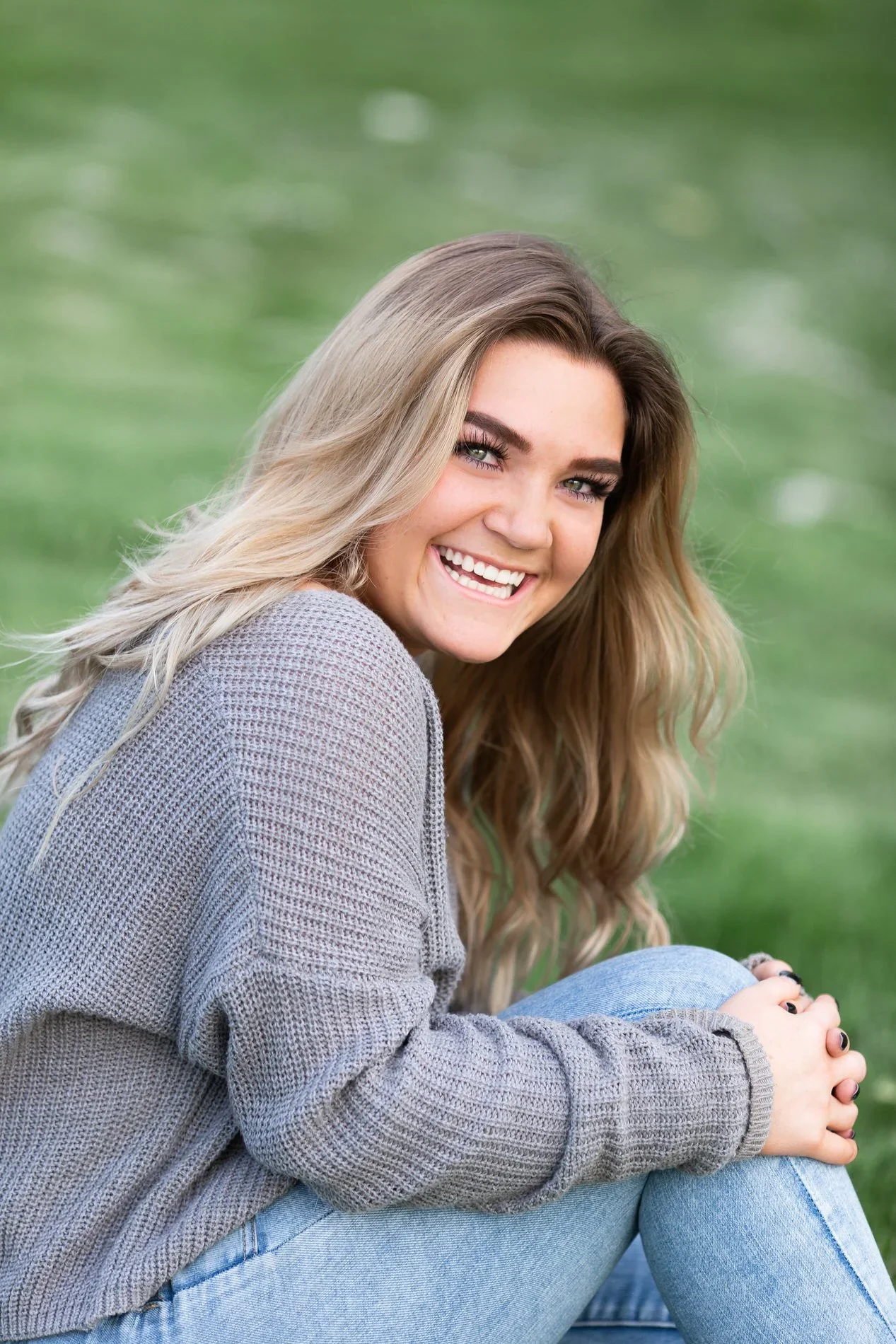 A smiling young woman with long, light brown hair, wearing a gray sweater and light blue jeans, sitting outdoors on grass.