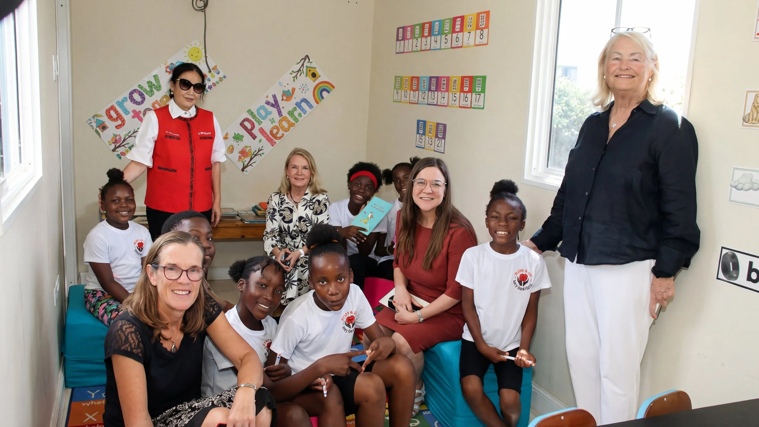A group of children and adults gathered in a classroom decorated with colorful educational posters and artwork, with some smiling and posing for the photo.