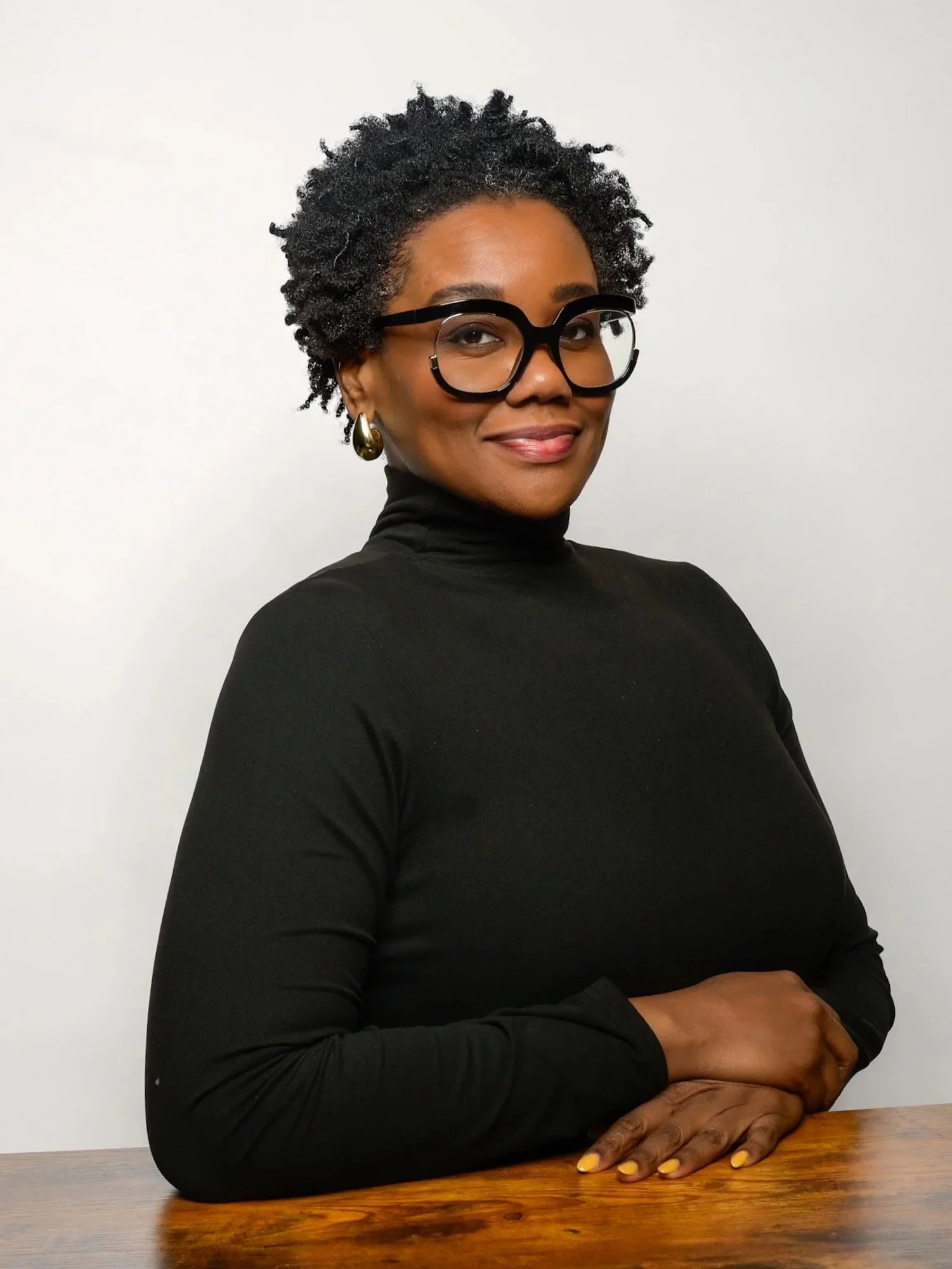 A woman with short curly black hair, wearing large black glasses, a black turtleneck, gold hoop earrings, and yellow nail polish, sitting at a wooden table against a plain white background.