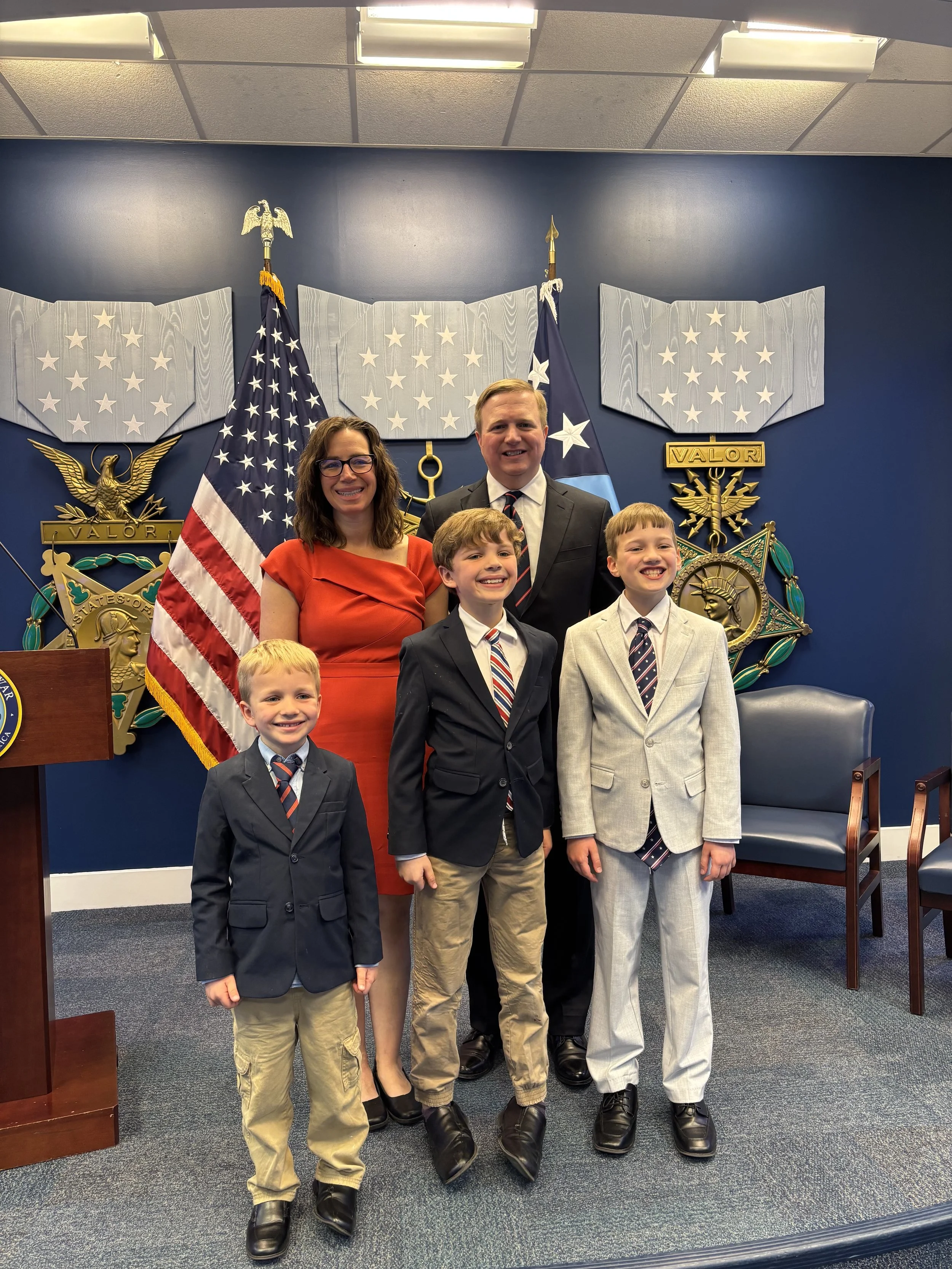 A group of five people, including two women and three young boys, standing together in a formal setting with American and state flags behind them. The background features a blue wall, military or official emblems, and a blue chair.