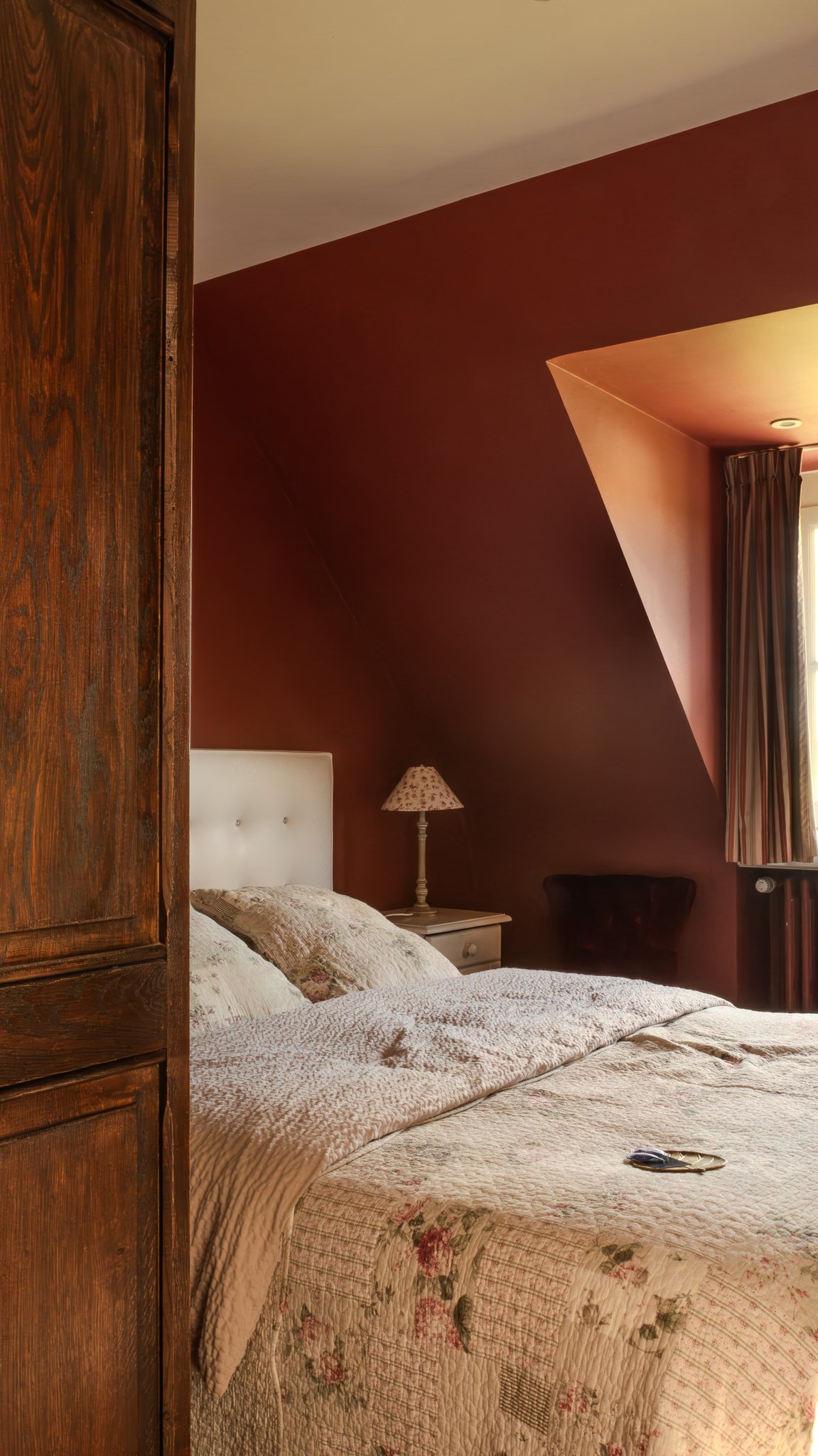 A cozy bedroom featuring a bed with a floral quilt, a white upholstered headboard, and a matching pillow. Next to the bed is a white nightstand with a lamp that has a floral lampshade. The room has a sloped ceiling painted in a warm reddish-brown tone, and a window with striped curtains allowing natural light. There's a dark armchair near the window and a large wooden wardrobe partially visible on the left.