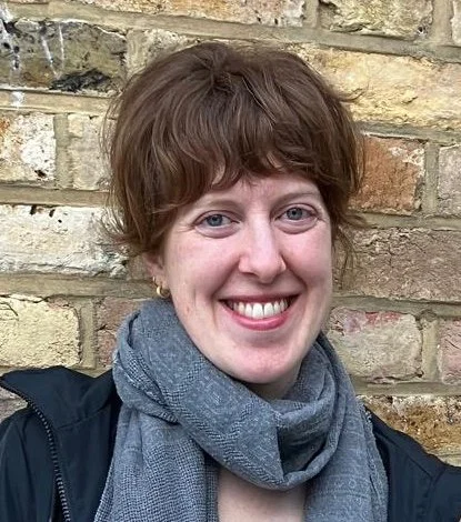 Woman with short brown hair smiling, wearing a gray scarf and black jacket, standing in front of a brick wall.