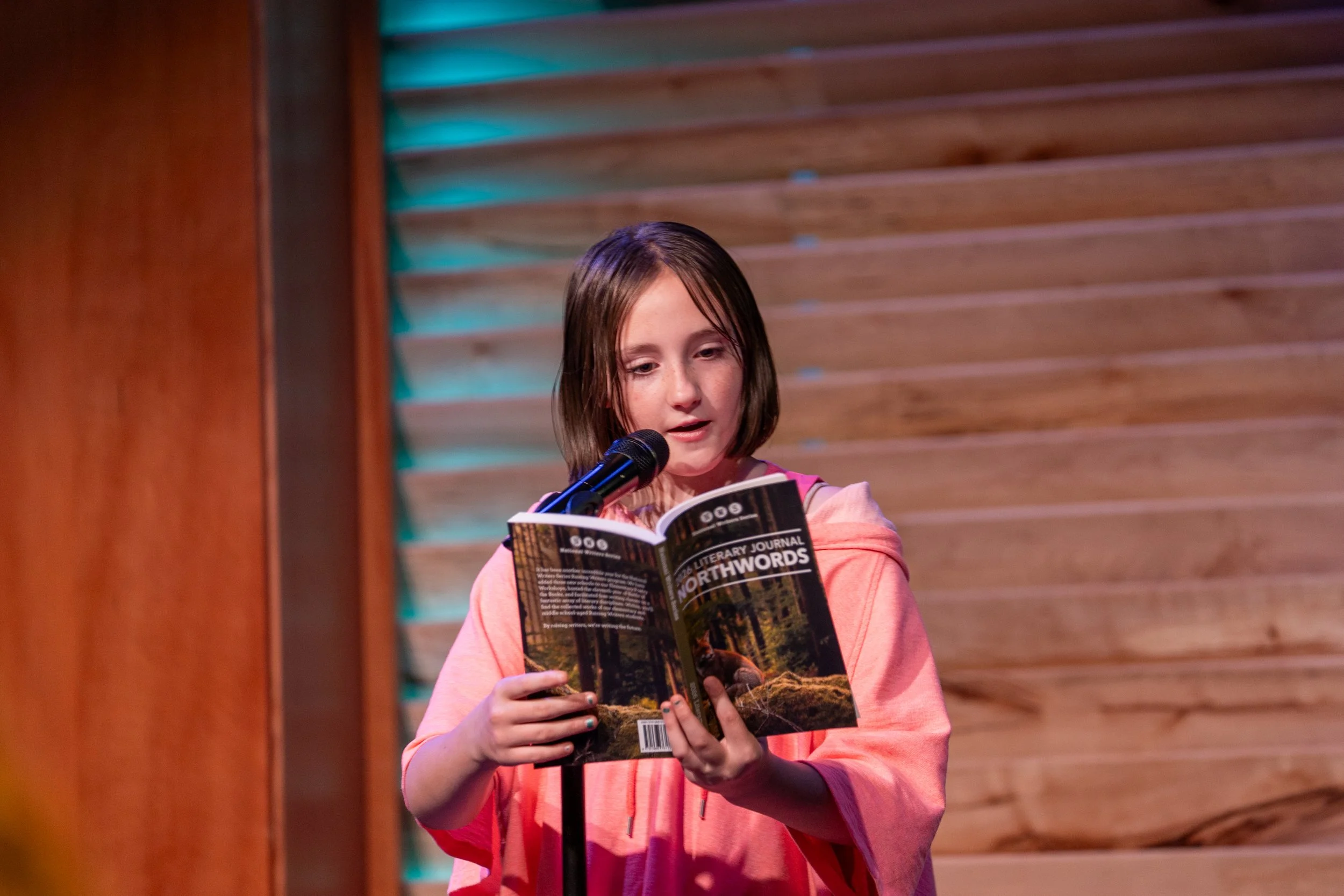 A young girl with shoulder-length brown hair and freckles reads a book titled "Northwords" into a microphone on a stand in front of a wooden wall with horizontal slats.
