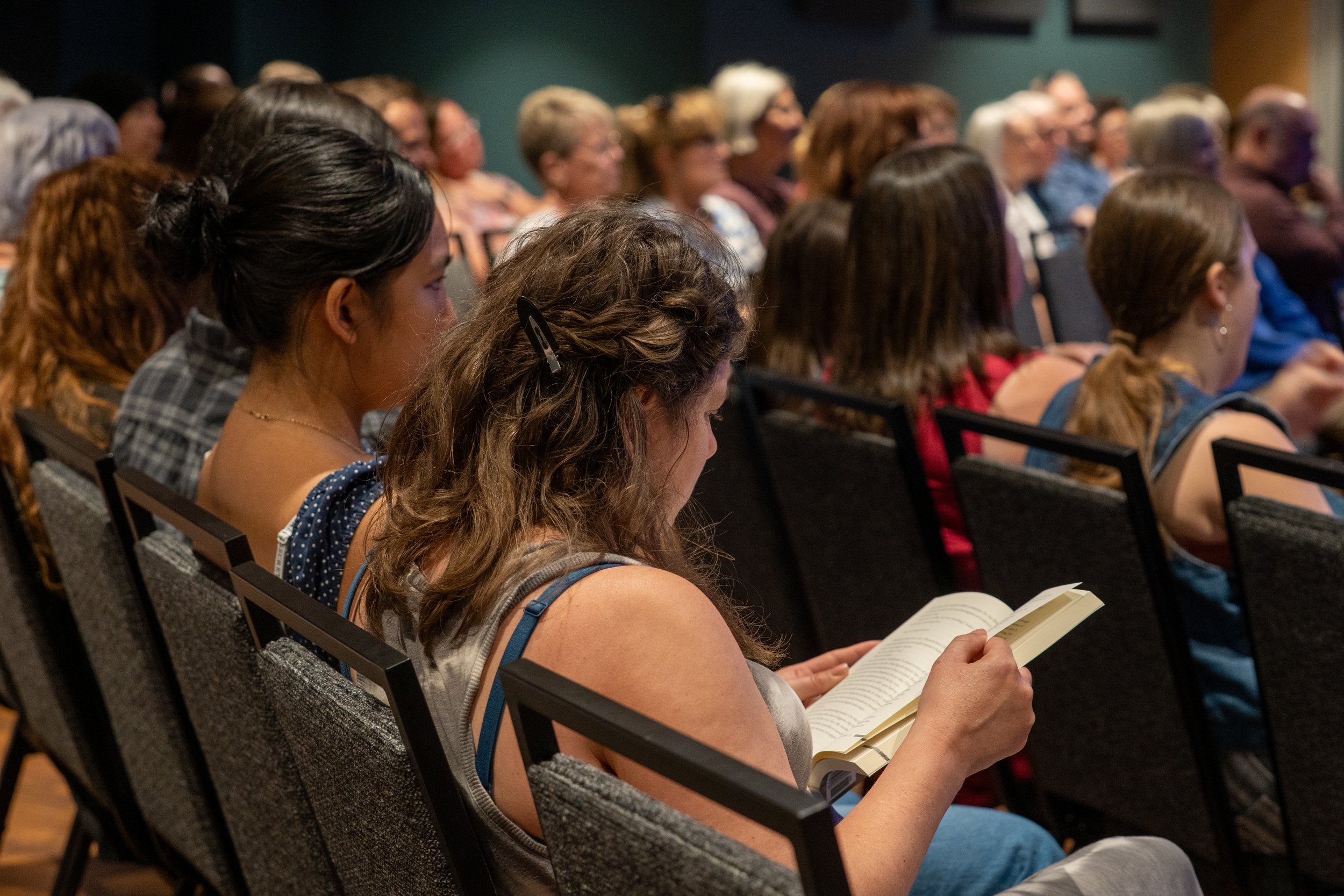 Audience attending a presentation or lecture, with attendees sitting in rows, some reading pamphlets or booklets, in an indoor setting.