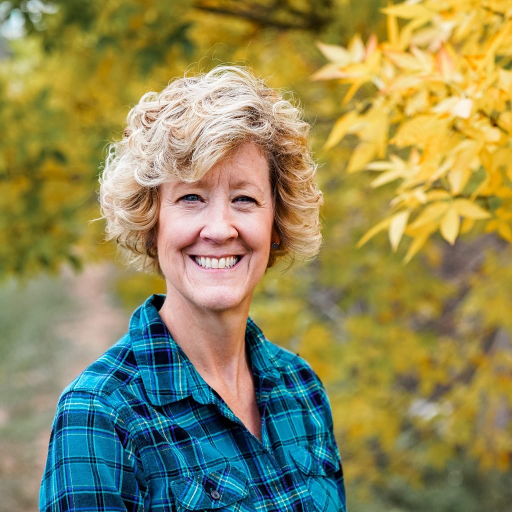 A woman with short curly blonde hair smiling outdoors in front of yellow autumn leaves, wearing a blue plaid shirt.