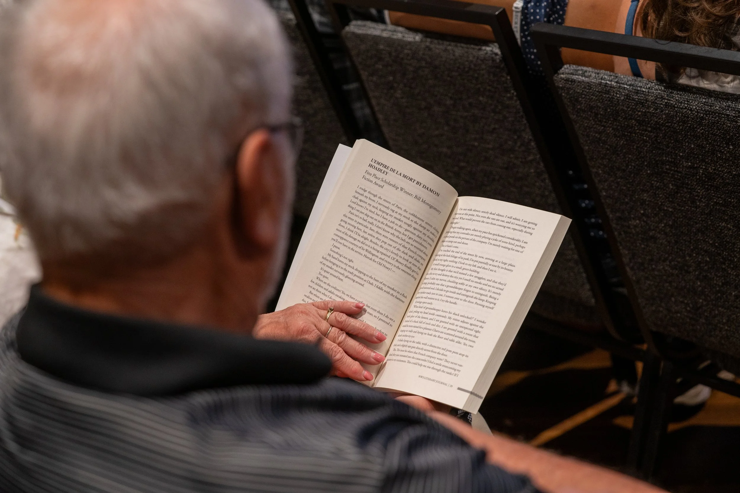 A person with gray hair reading a book in a seated position, sitting outdoors or in a public space, with other people seated nearby and chairs with fabric seats.