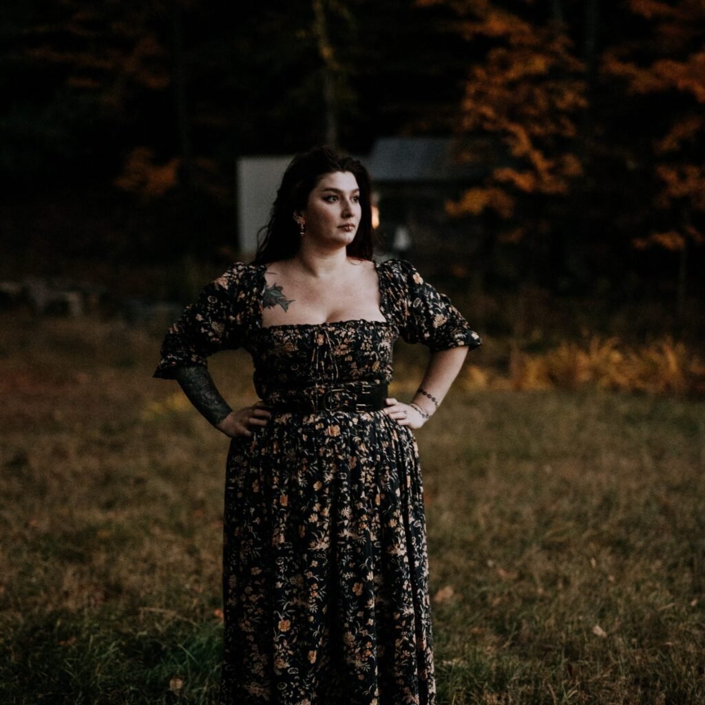 A woman with dark hair standing outdoors at dusk, wearing a long black floral dress with puffed sleeves, with hands on her hips, in a grassy area with trees and a building in the background.