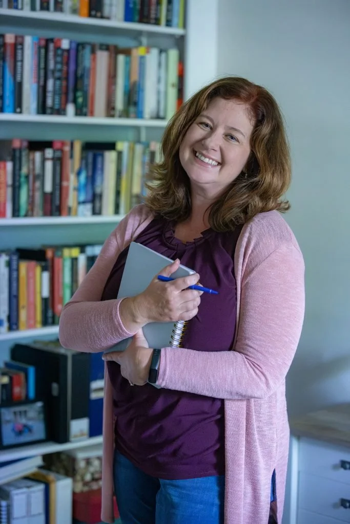 A woman with shoulder-length red hair, smiling, holding a notebook and pen, standing in front of a bookshelf filled with books.