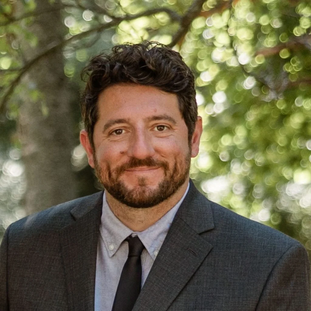 A man with dark curly hair and a beard, wearing a gray suit, light blue shirt, and black tie, standing outdoors with trees and green foliage in the background.