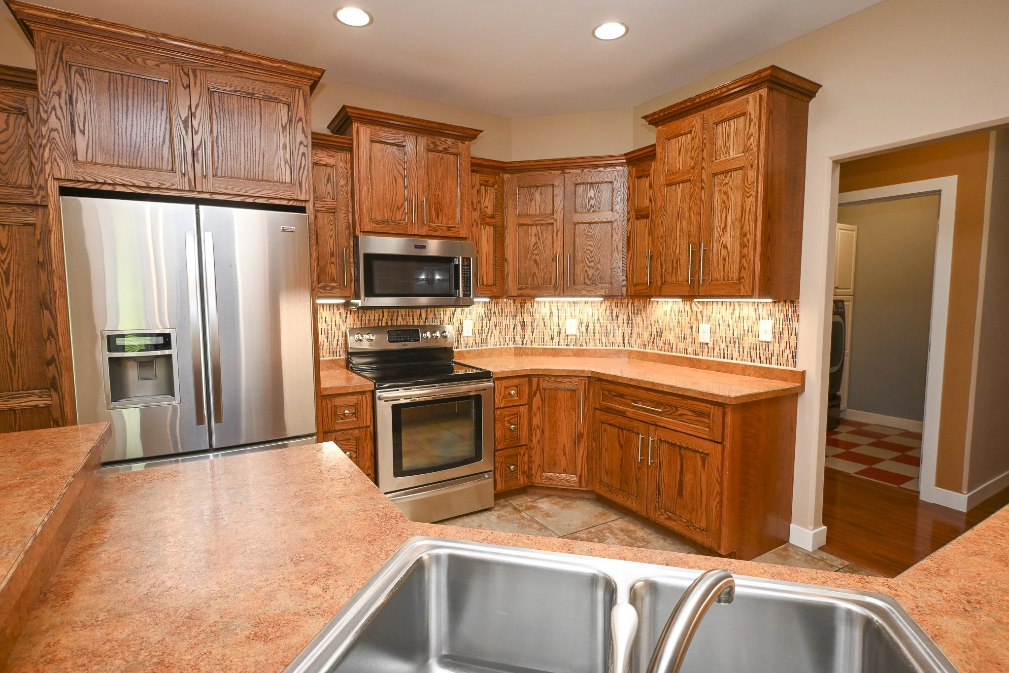 Kitchen with wooden cabinets, stainless steel refrigerator, microwave, and an oven. Colorful tile backsplash, pinkish countertop, and a stainless steel sink in the foreground.