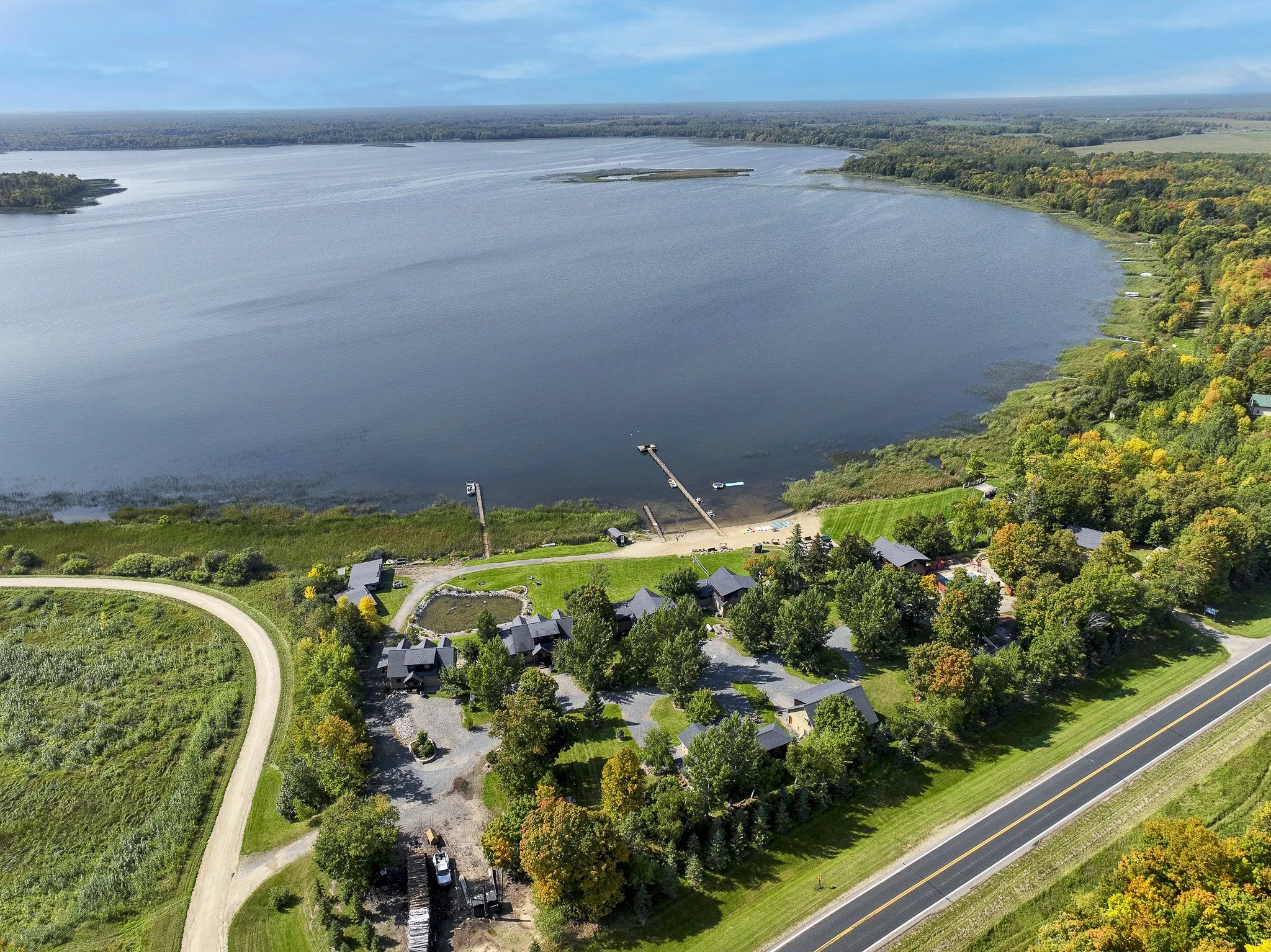 Aerial view of a lake with surrounding greenery, trees, and houses, with a boat dock and boat on the water, and a winding road along the shoreline.