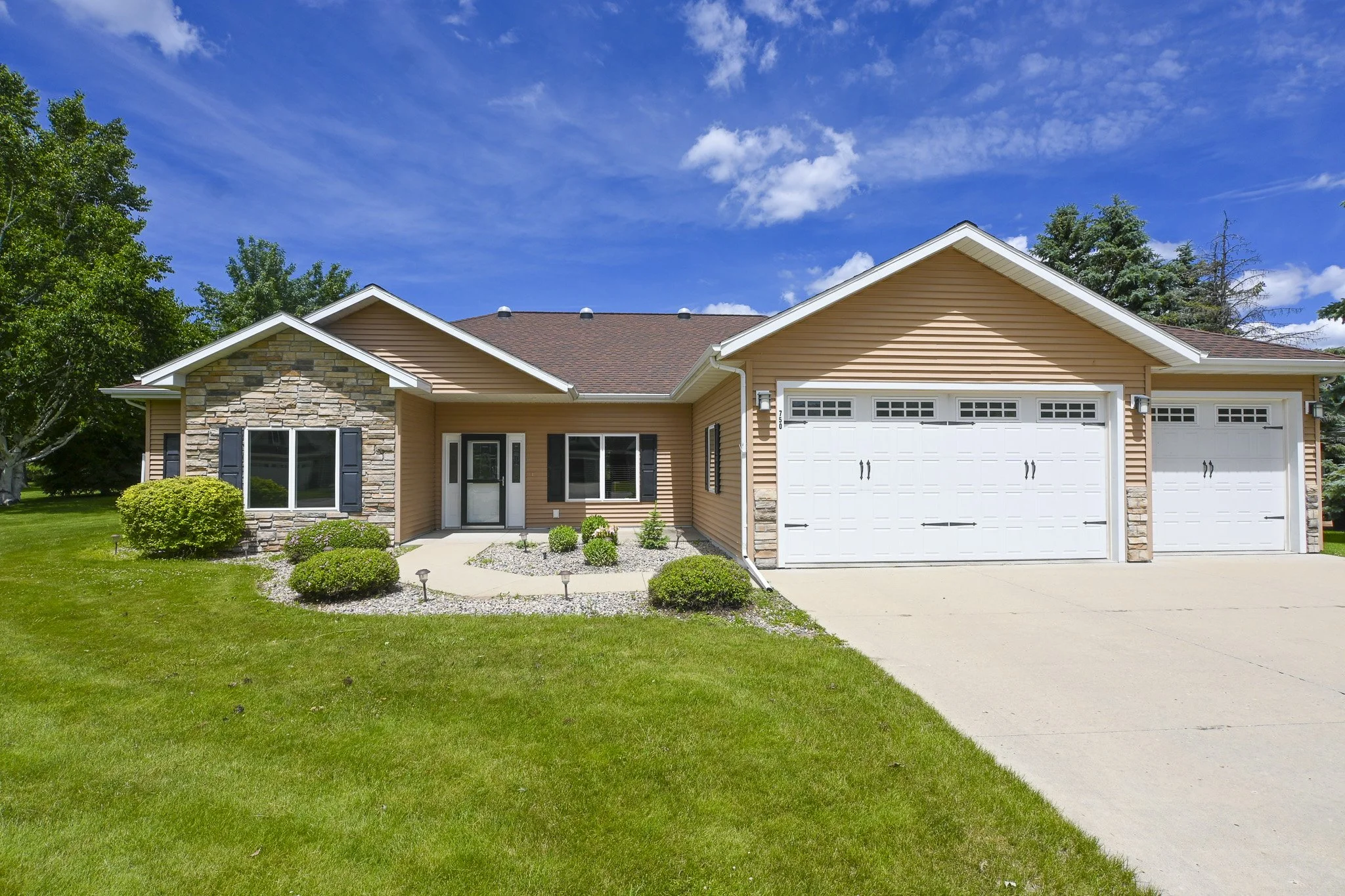 Front view of a suburban house with beige siding, stone accents, a two-car garage, green lawn, and a blue sky with clouds.