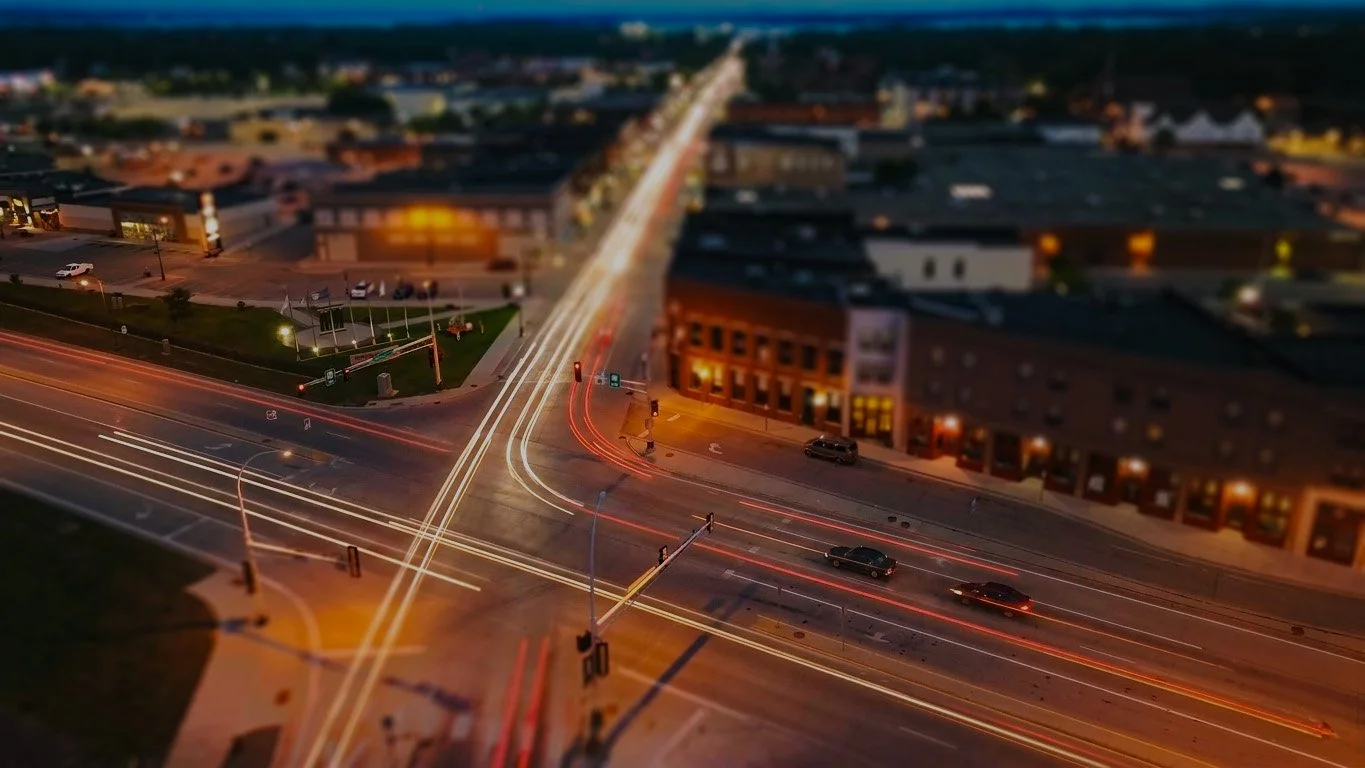 Long exposure photo of a city street intersection at dusk with light trails from moving vehicles, buildings, streetlights, and traffic signals.
