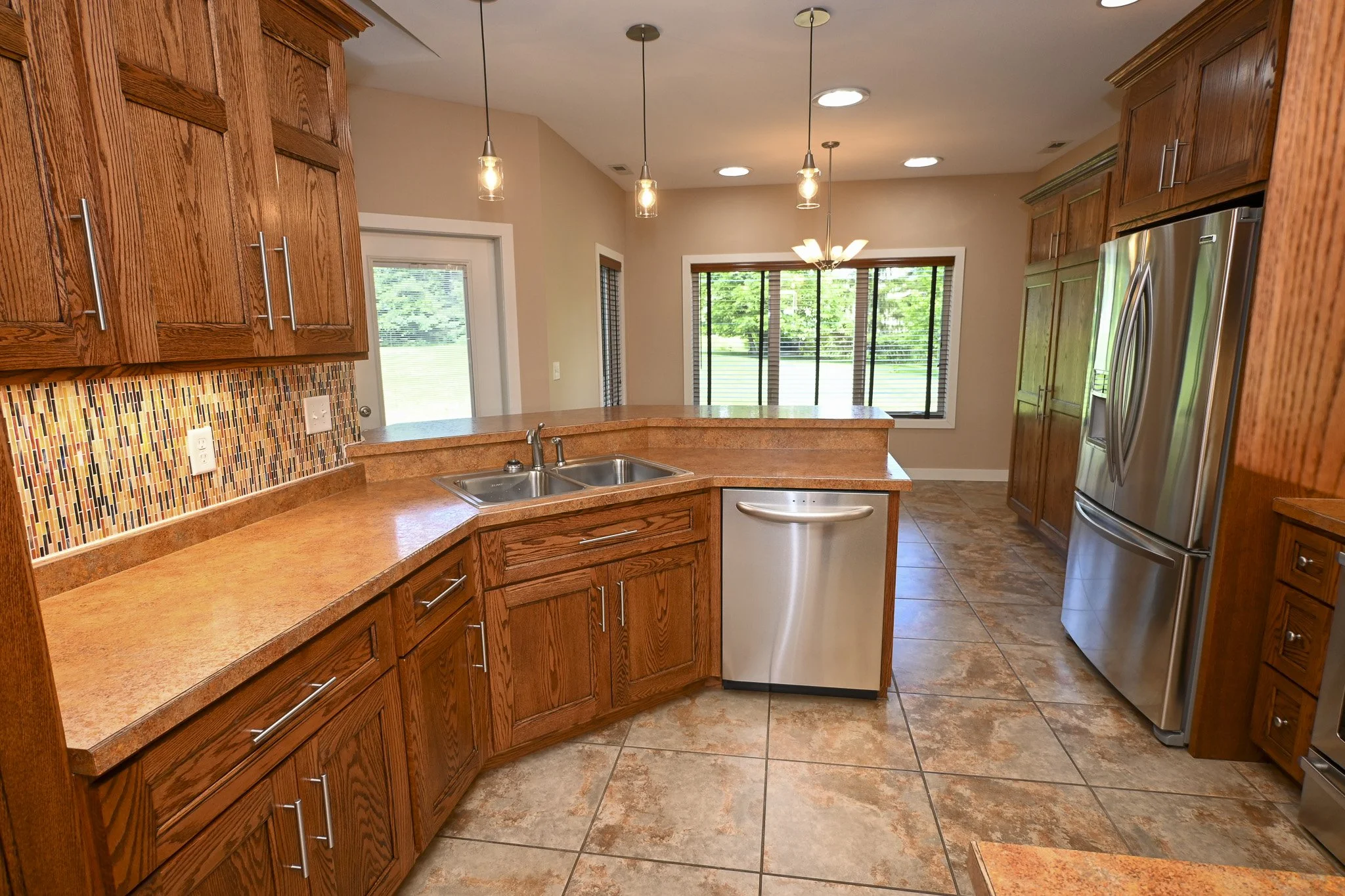 Kitchen with wooden cabinets, a double sink, a stainless steel dishwasher, a side-by-side refrigerator, tiled floor, pendant lights and large windows showing green outdoor scenery.