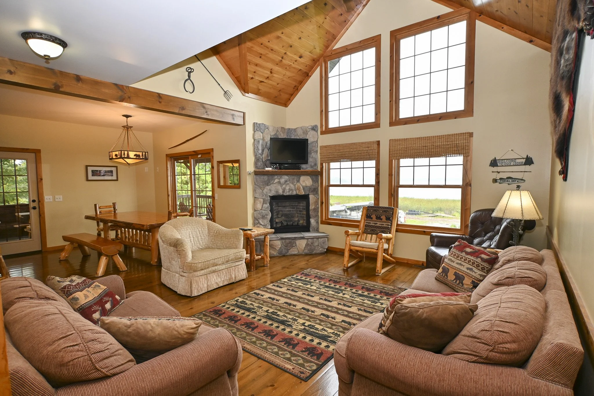 Living room with high wooden vaulted ceiling, large windows, stone fireplace with a TV, and rustic furniture including sofas and chairs.