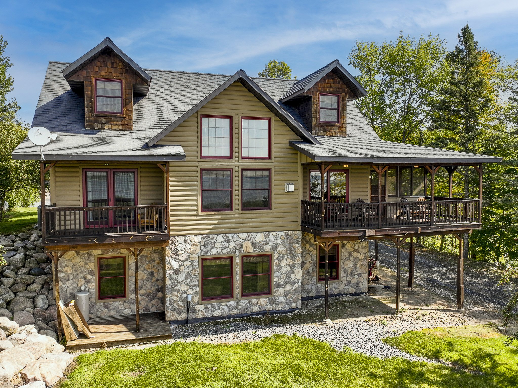 A two-story house with beige siding, stone foundation, and a gabled roof with gray shingles. The house has multiple windows with red frames and a large wraparound porch. The house is surrounded by trees and a grassy yard, with some construction work ongoing at the deck area.