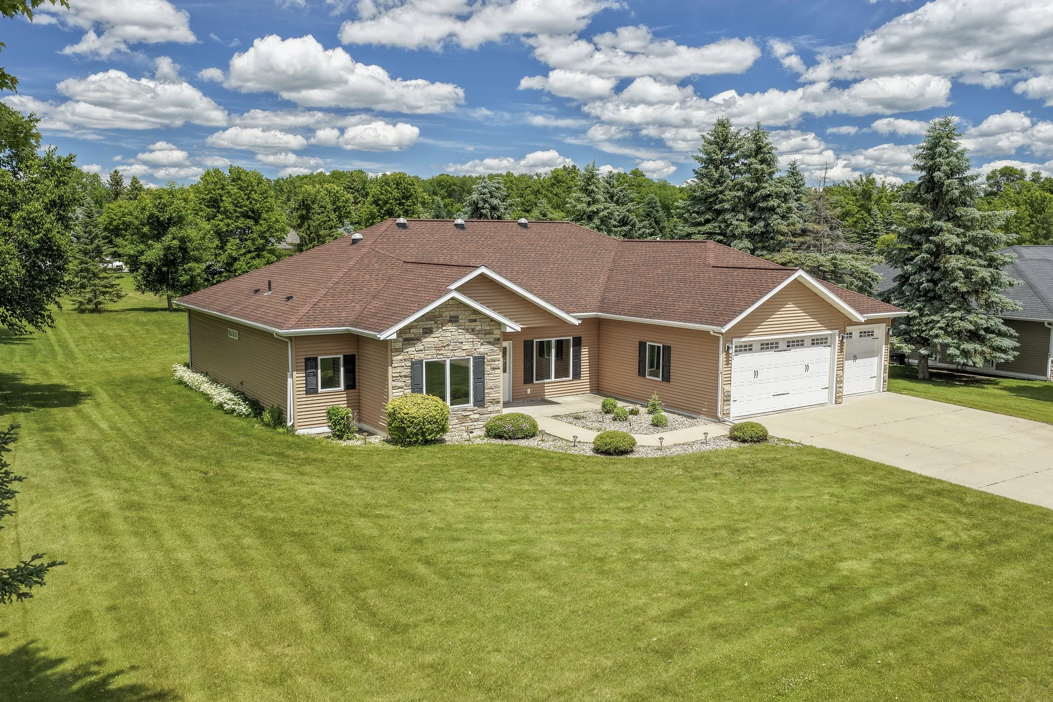 A house with a brown roof and beige siding, surrounded by a green lawn and trees, under a partly cloudy sky.