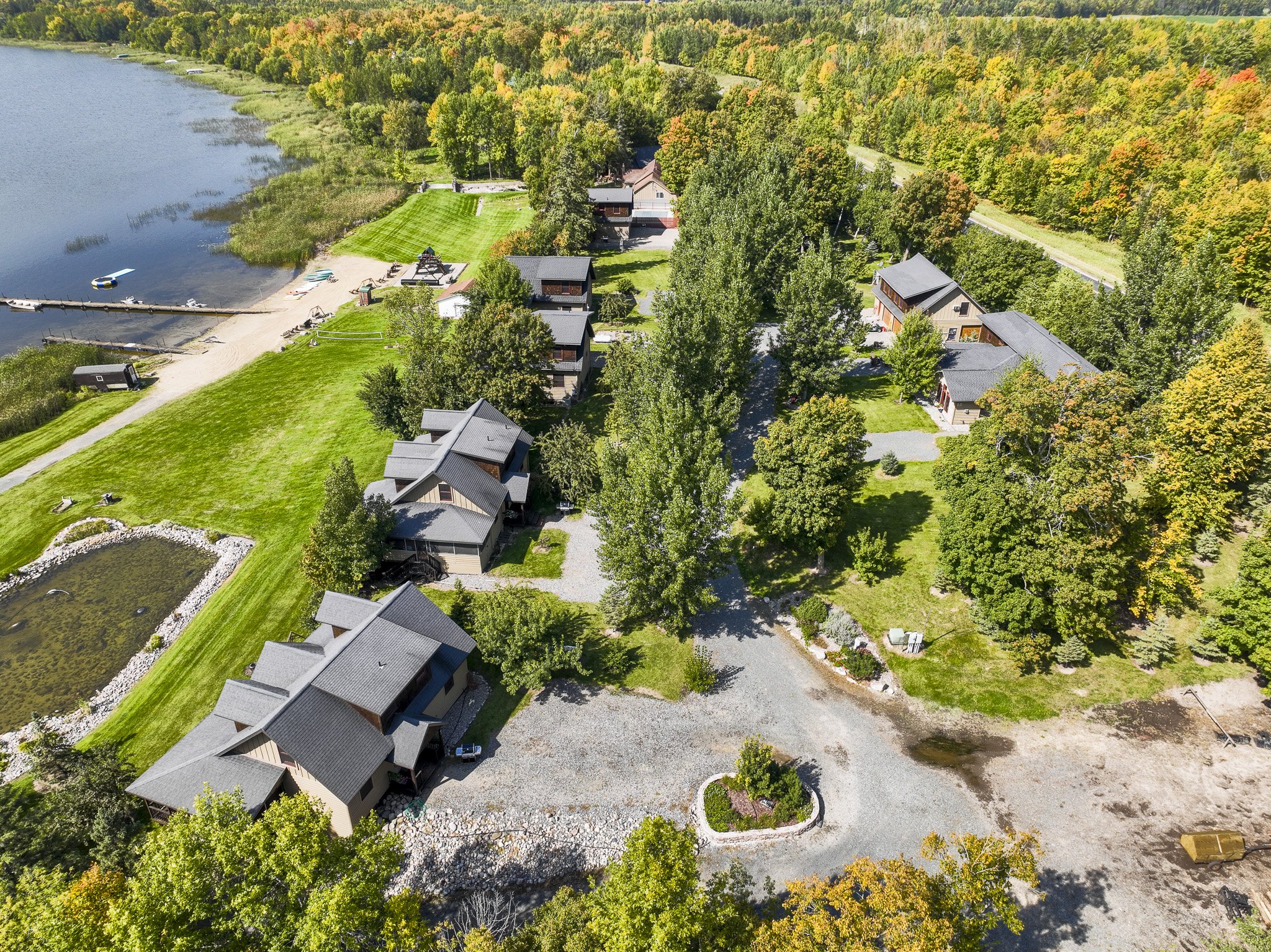 Aerial view of a lakeside residential area with several houses, lush green trees, a small beach, and a dock on the water, surrounded by dense forest with fall foliage.