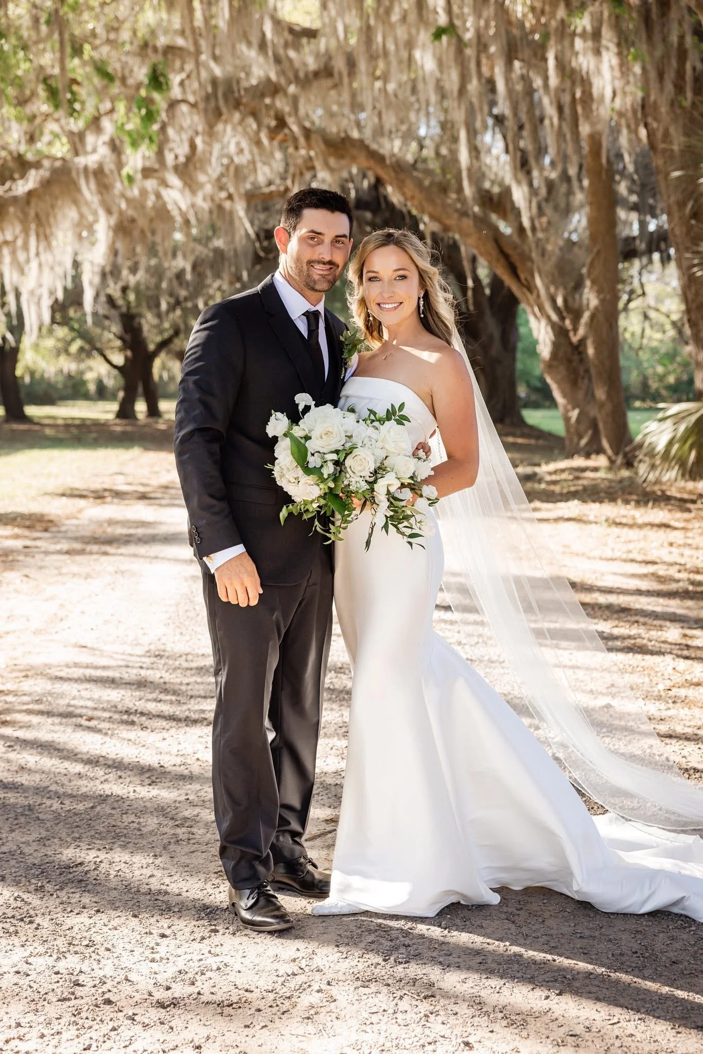 A happy bride and groom standing outdoors with trees in the background. The groom wears a black suit and tie, and the bride wears a white wedding dress with a veil. She holds a bouquet of white flowers with green leaves.