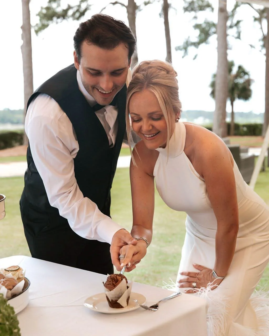 A bride and groom cutting and serving cake at their wedding reception outdoors, surrounded by trees and a scenic view.