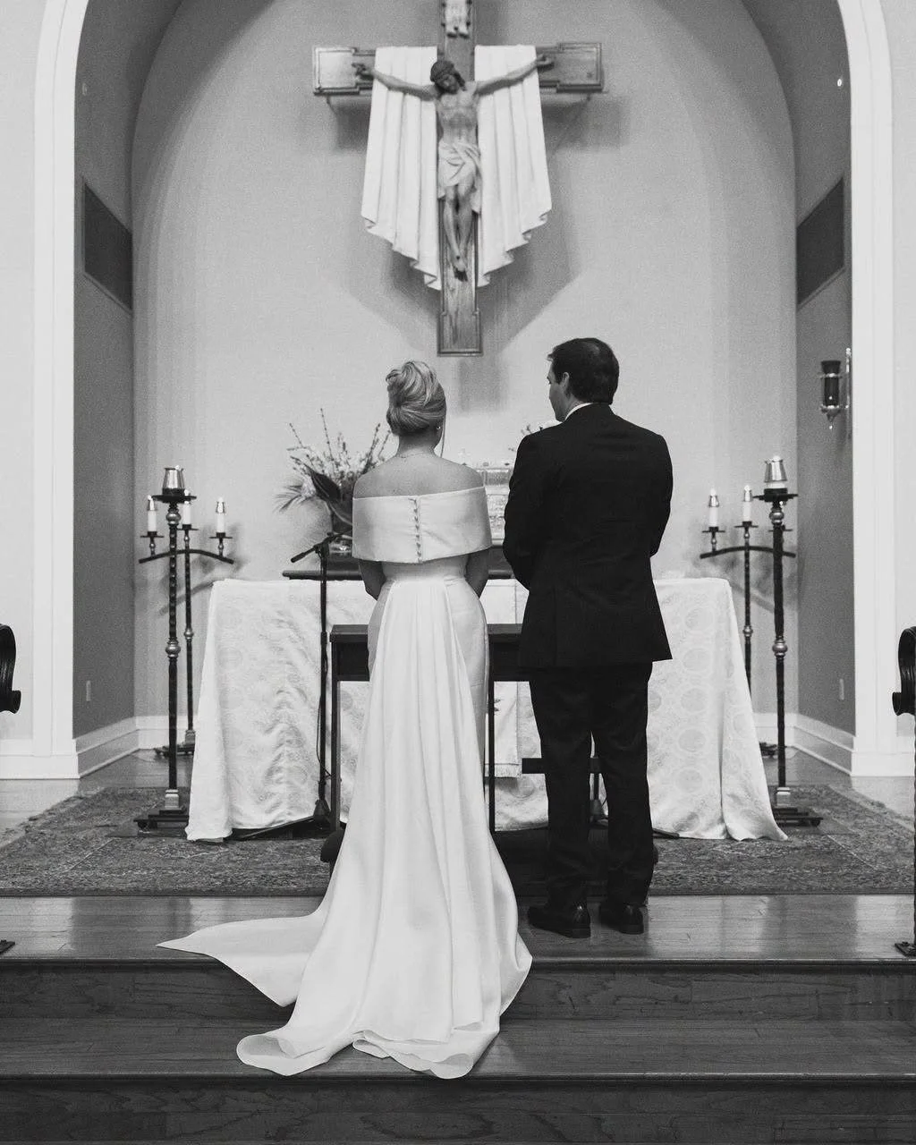 A black and white photo of a wedding ceremony in a church, showing a bride and groom standing before an altar with a crucifix above, facing away from the camera.
