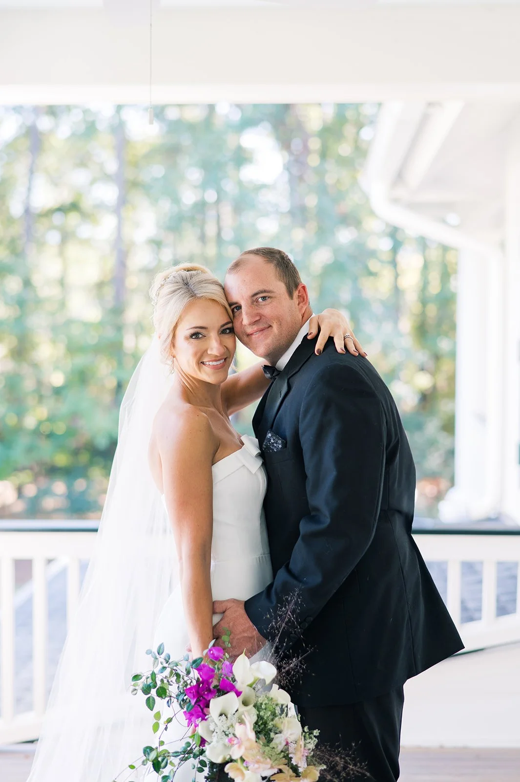 A bride and groom on their wedding day smiling and embracing, with a bouquet of flowers. The bride is wearing a white strapless wedding gown and veil, and the groom is dressed in a black tuxedo, outdoors with trees in the background.