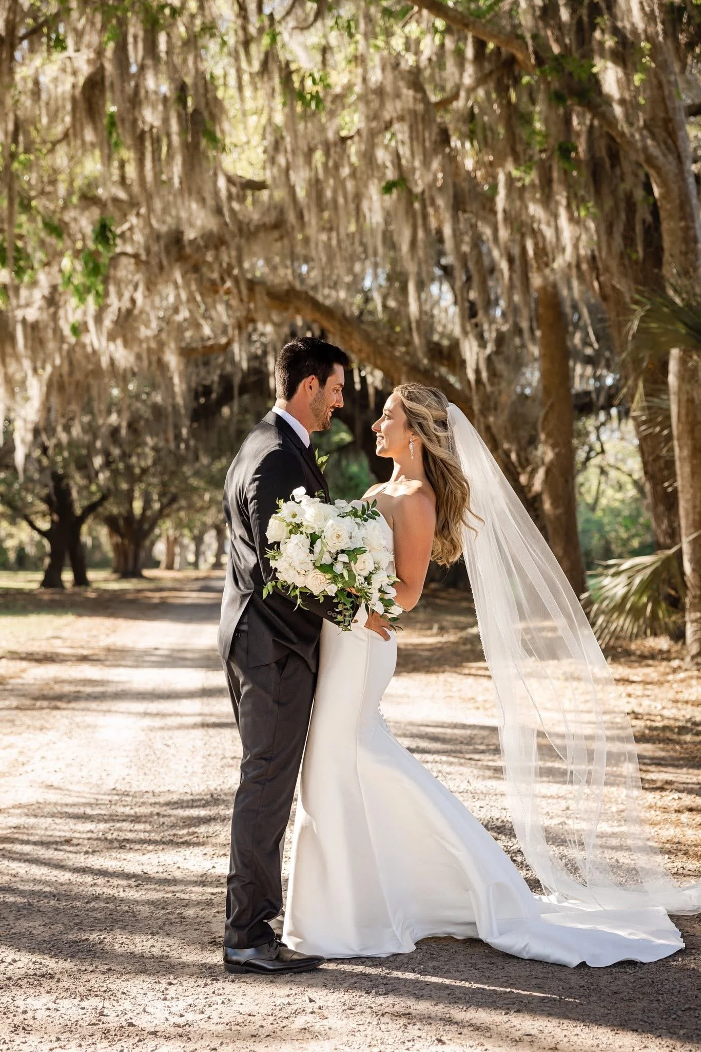 A bride and groom standing face to face under a canopy of trees, smiling at each other, with the bride holding a bouquet of white flowers, during a wedding ceremony outdoors.