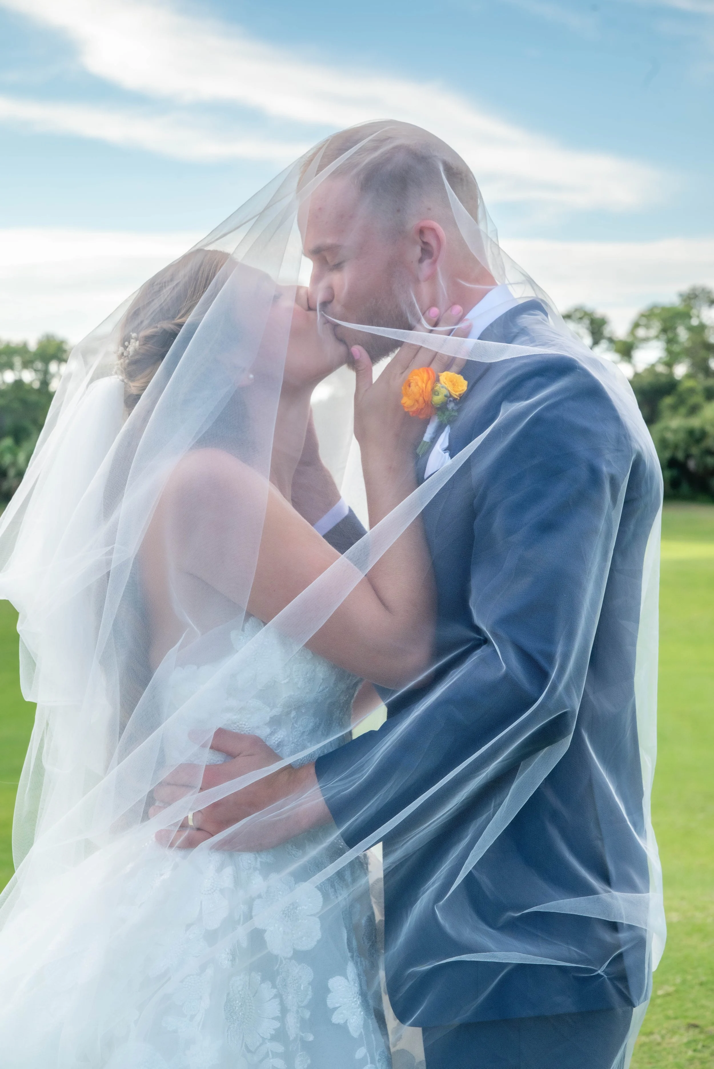 A newlywed couple kissing under a wedding veil outdoors with a grassy field and blue sky in the background.