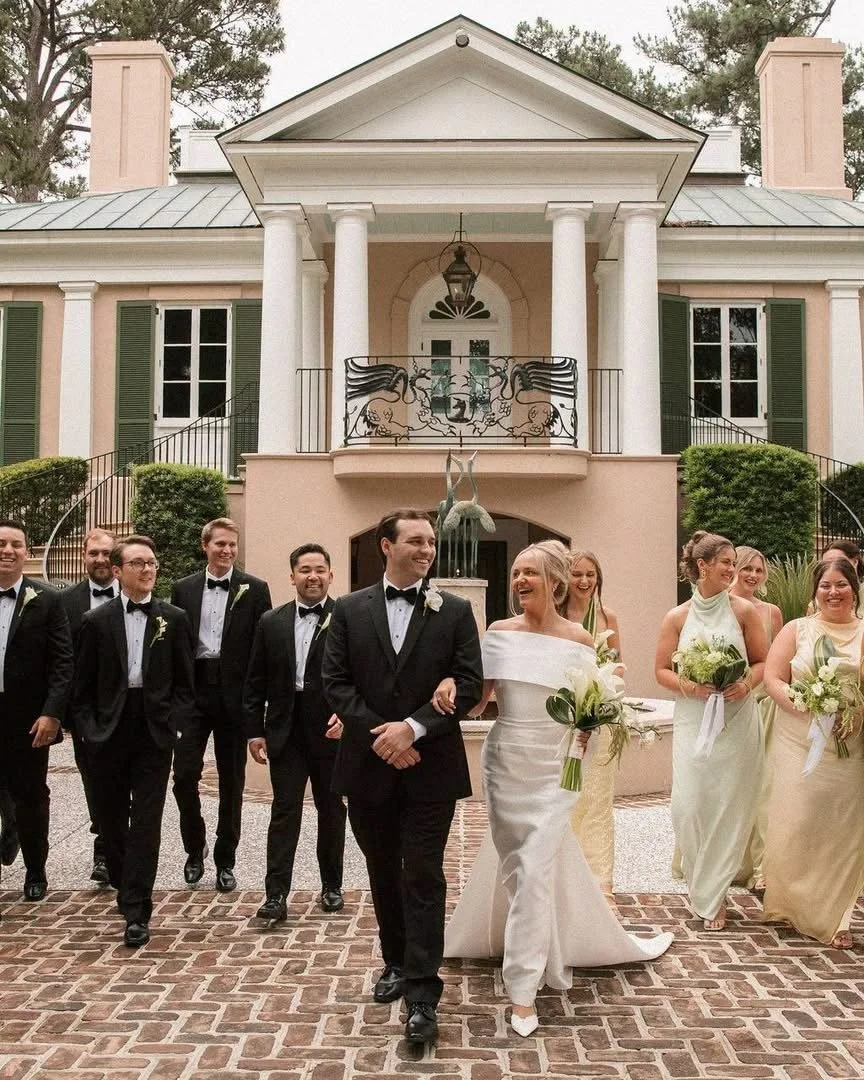 Wedding party walking in front of a large, elegant house with columns and green shutters.
