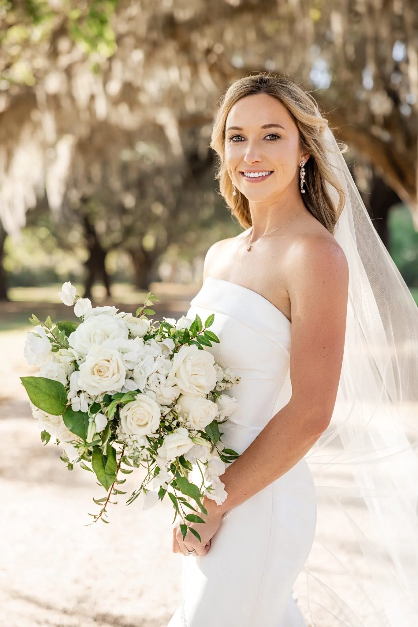 A smiling bride with light skin and blonde hair, wearing a strapless white wedding dress and dangling earrings, holding a large bouquet of white roses and greenery outdoors with trees in the background.