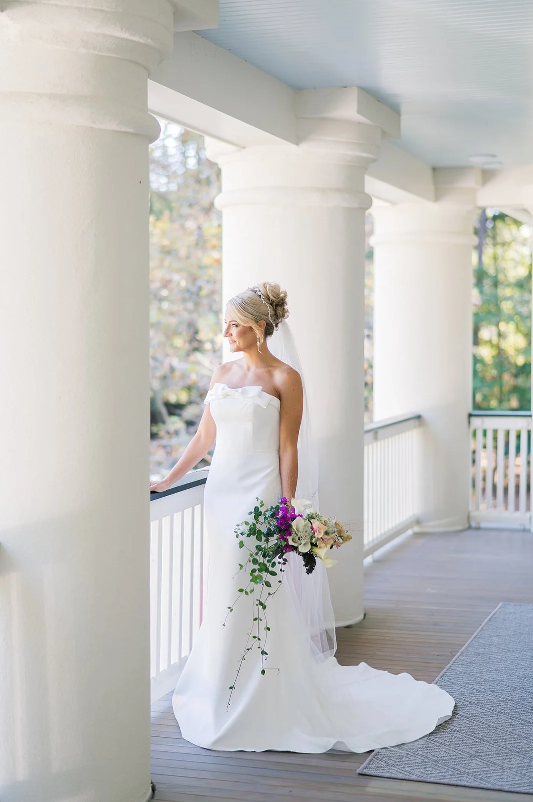 A bride in a white wedding gown holding a bouquet of flowers on a porch with white columns and railings.