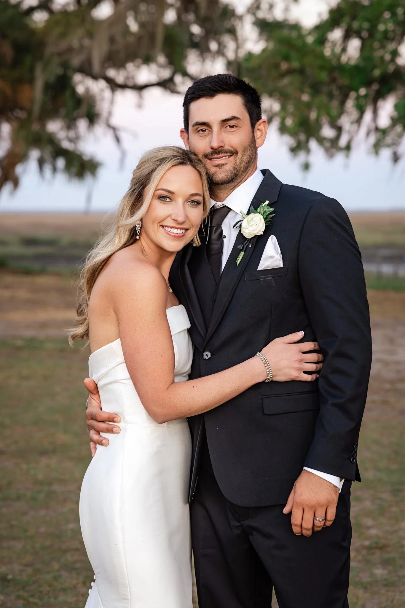 A happy bride and groom standing outdoors, embracing and smiling. The bride is in a white wedding dress and the groom is in a black tuxedo with a white boutonniere. Background of trees and soft sky.