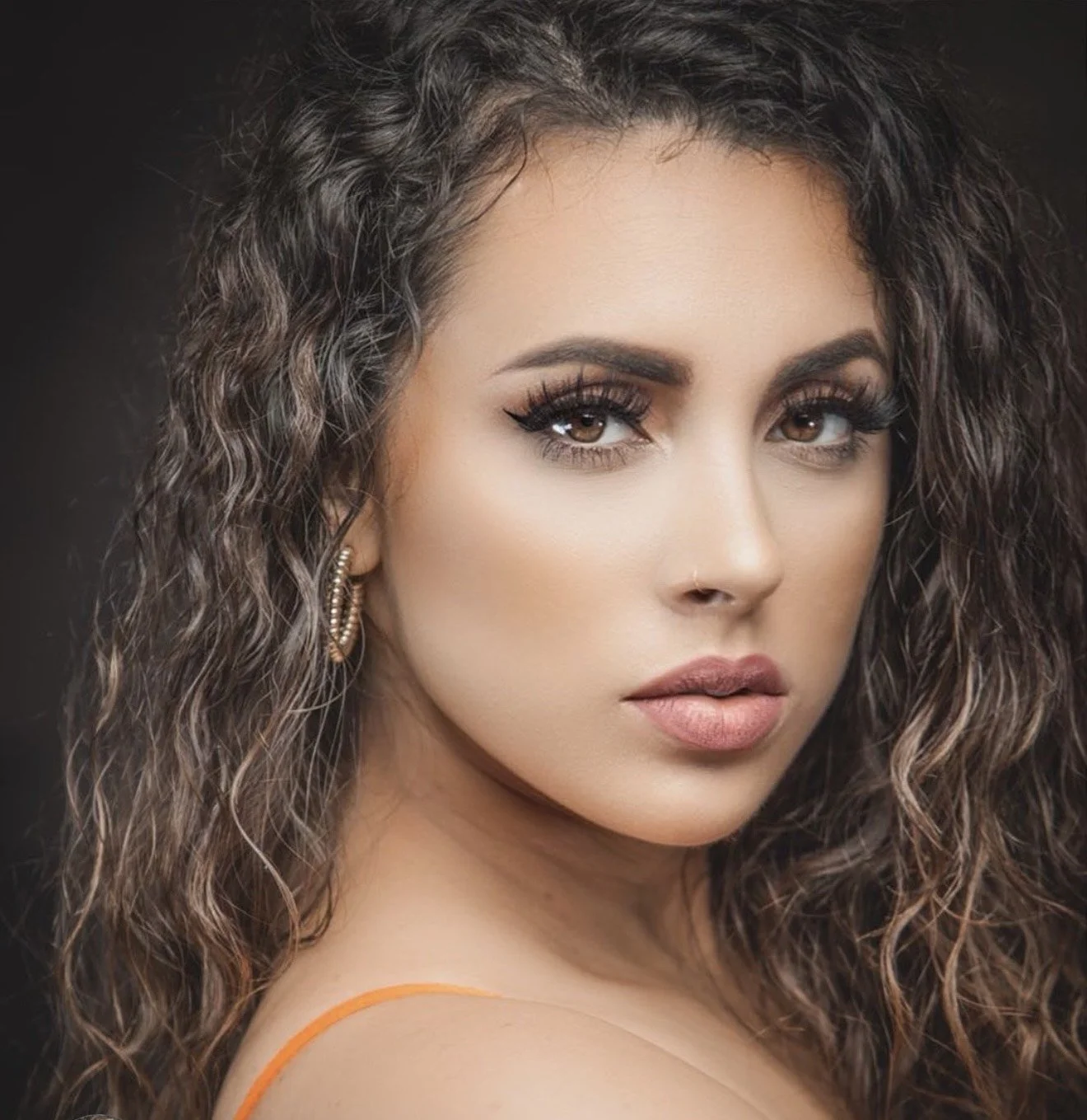 Close-up portrait of a woman with curly brown hair, makeup, and gold earrings, posing against a dark background.