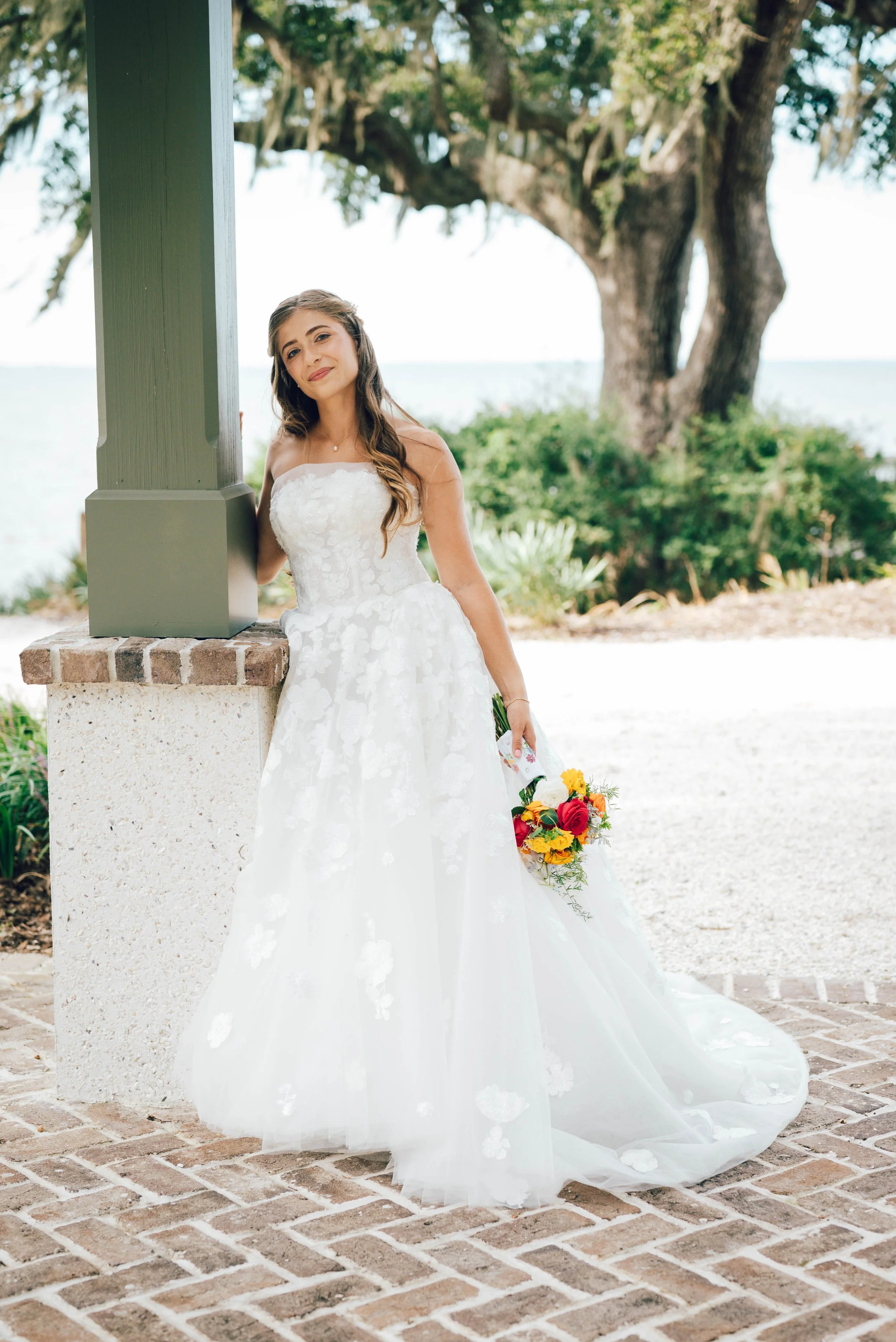 A bride in a white wedding gown holding a colorful bouquet, standing outdoors near a large tree and a green lamppost.