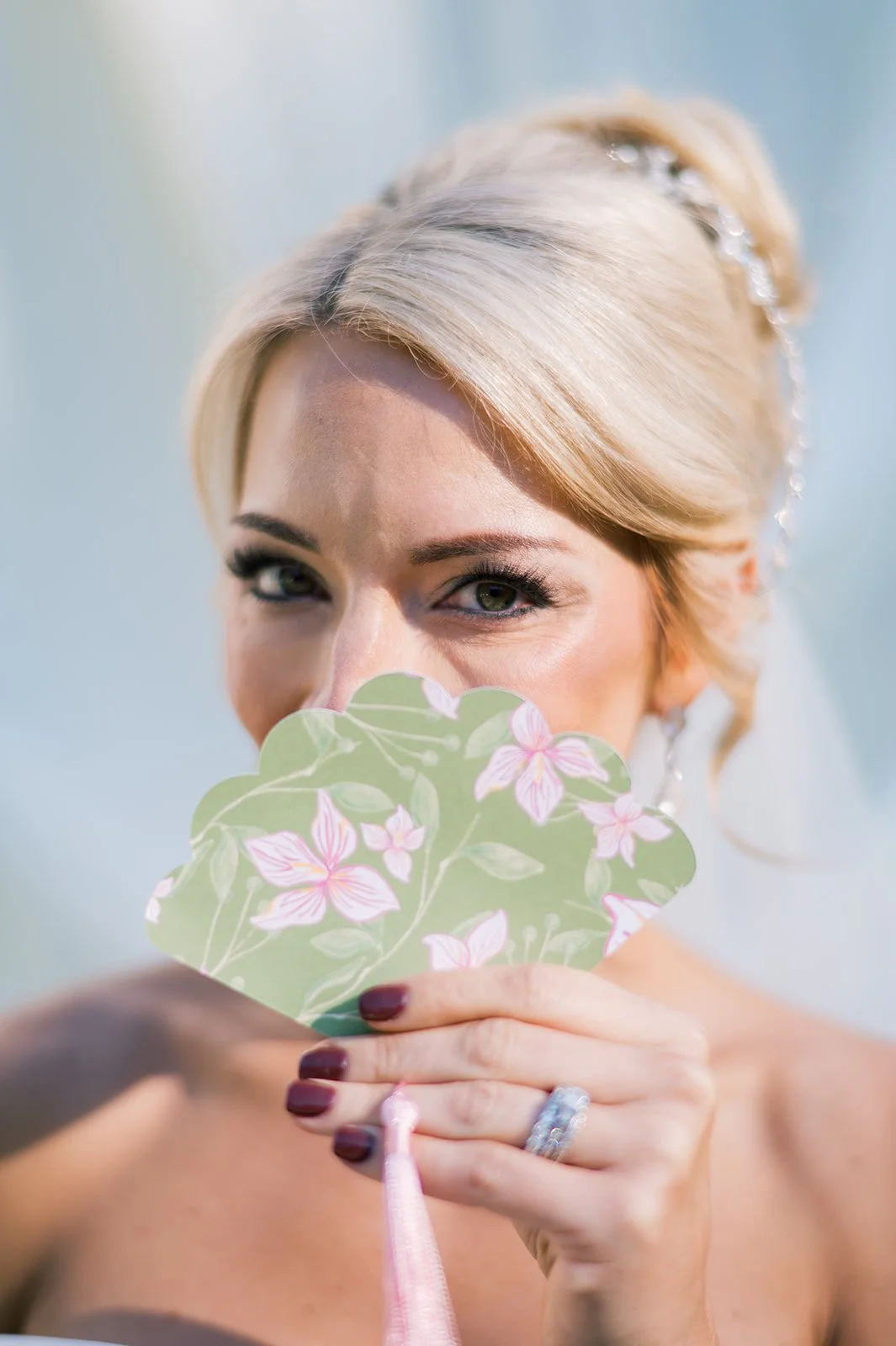 A woman with blonde hair, partially covering her face with a floral fan, looking at the camera with a wink.