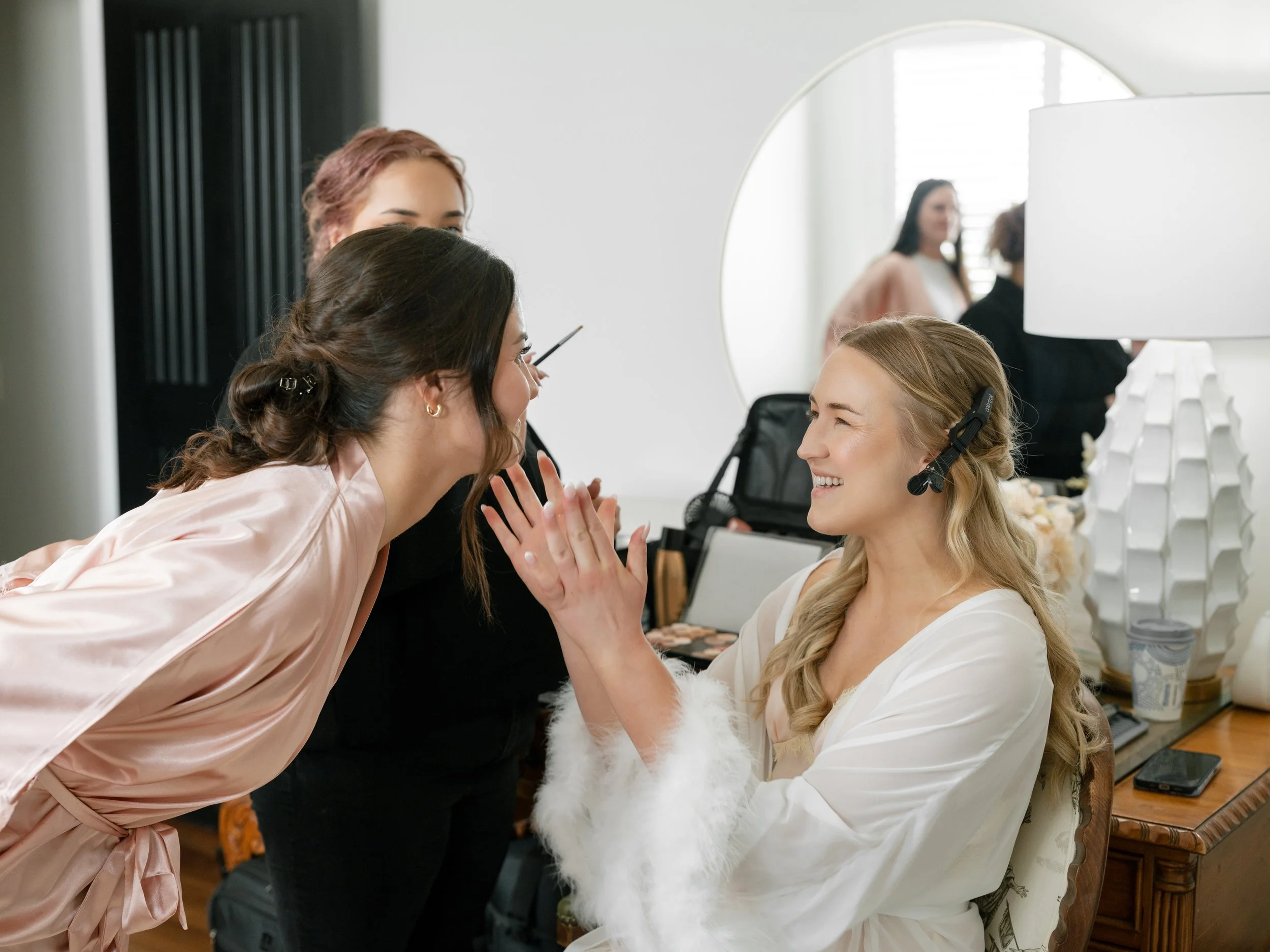 A bride and a woman with long brown hair, holding her hands and smiling, are engaging in a friendly conversation at a bridal preparation area. Other women and makeup artists are visible in the background.