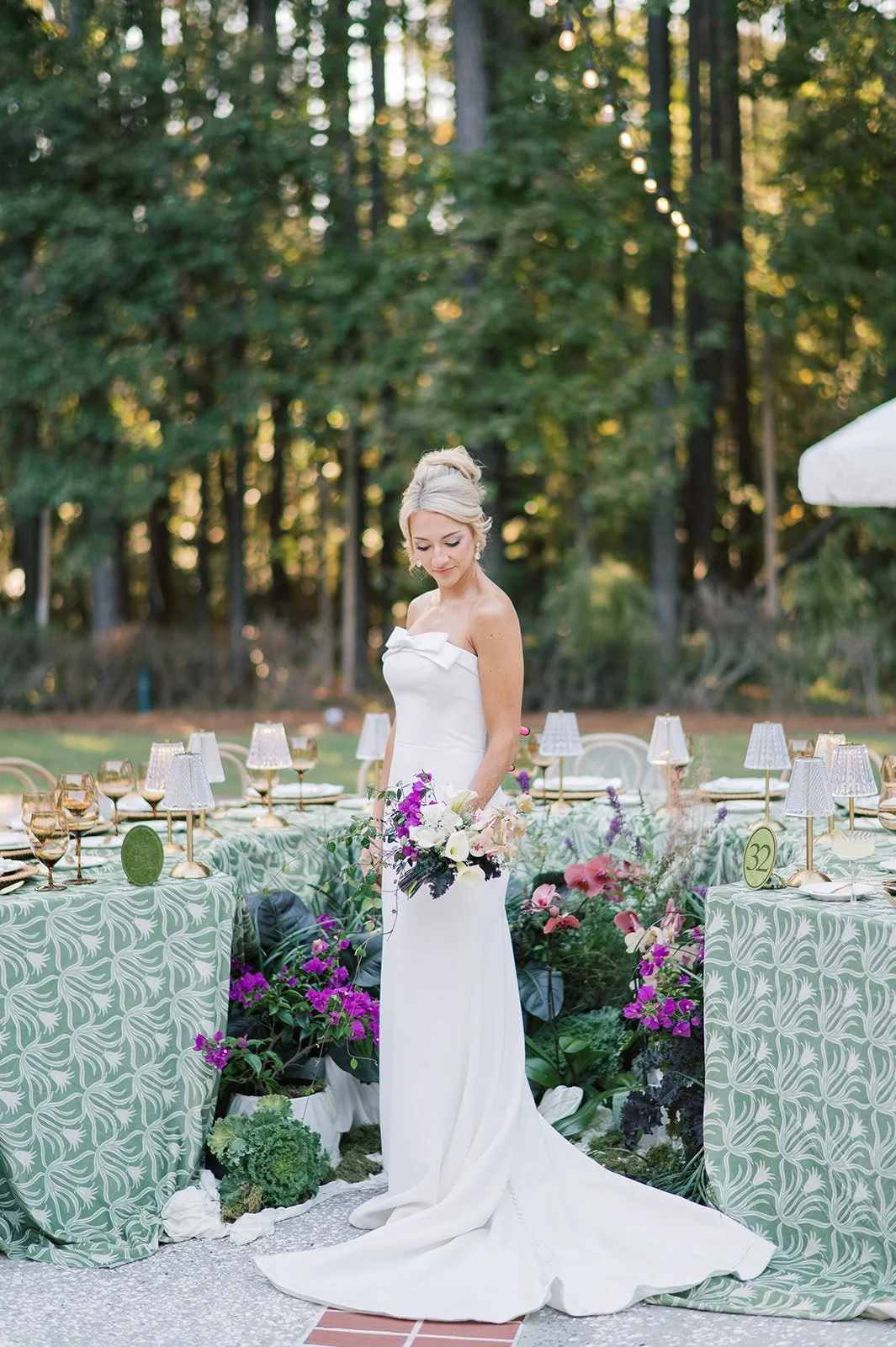 A bride in a white wedding gown holding a bouquet of flowers stands outdoors at a decorated reception table with patterned green tablecloths, flower arrangements, and table lamps, in a wooded area during daylight.