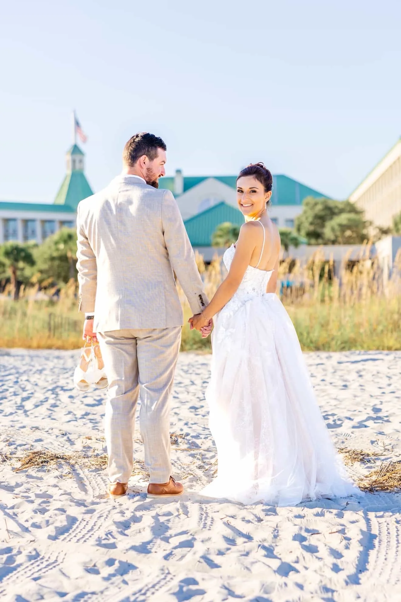 A bride and groom holding hands on a sandy beach, smiling at the camera with a large building with green roofs in the background. The bride is wearing a white wedding gown, and the groom is dressed in a light-colored suit. The sky is clear and sunny.