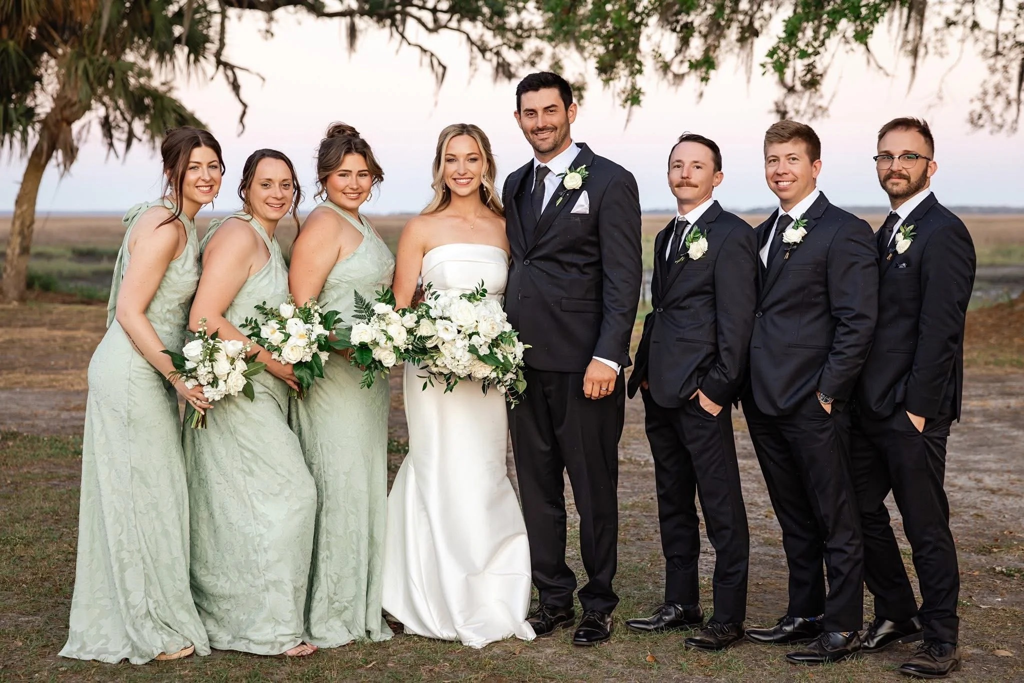 Group of wedding guests, including the bride and groom, standing outdoors with trees in the background.