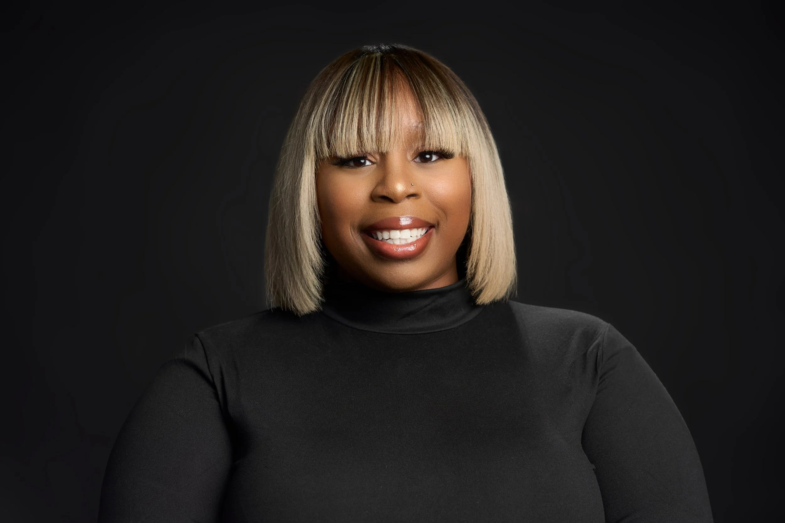 A portrait of a woman with straight blonde hair with bangs, wearing a black top, smiling against a dark background.