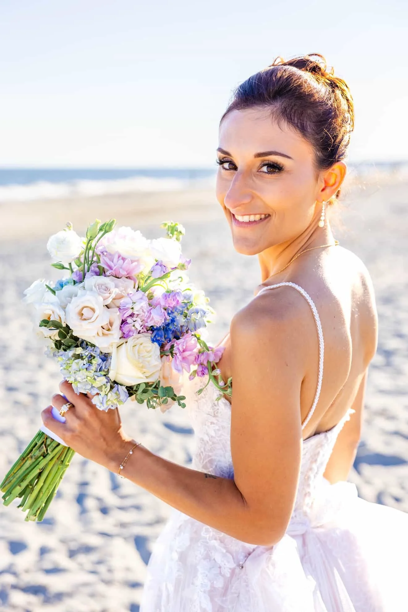 A smiling bride holding a colorful bouquet of flowers on a sandy beach with the ocean in the background.