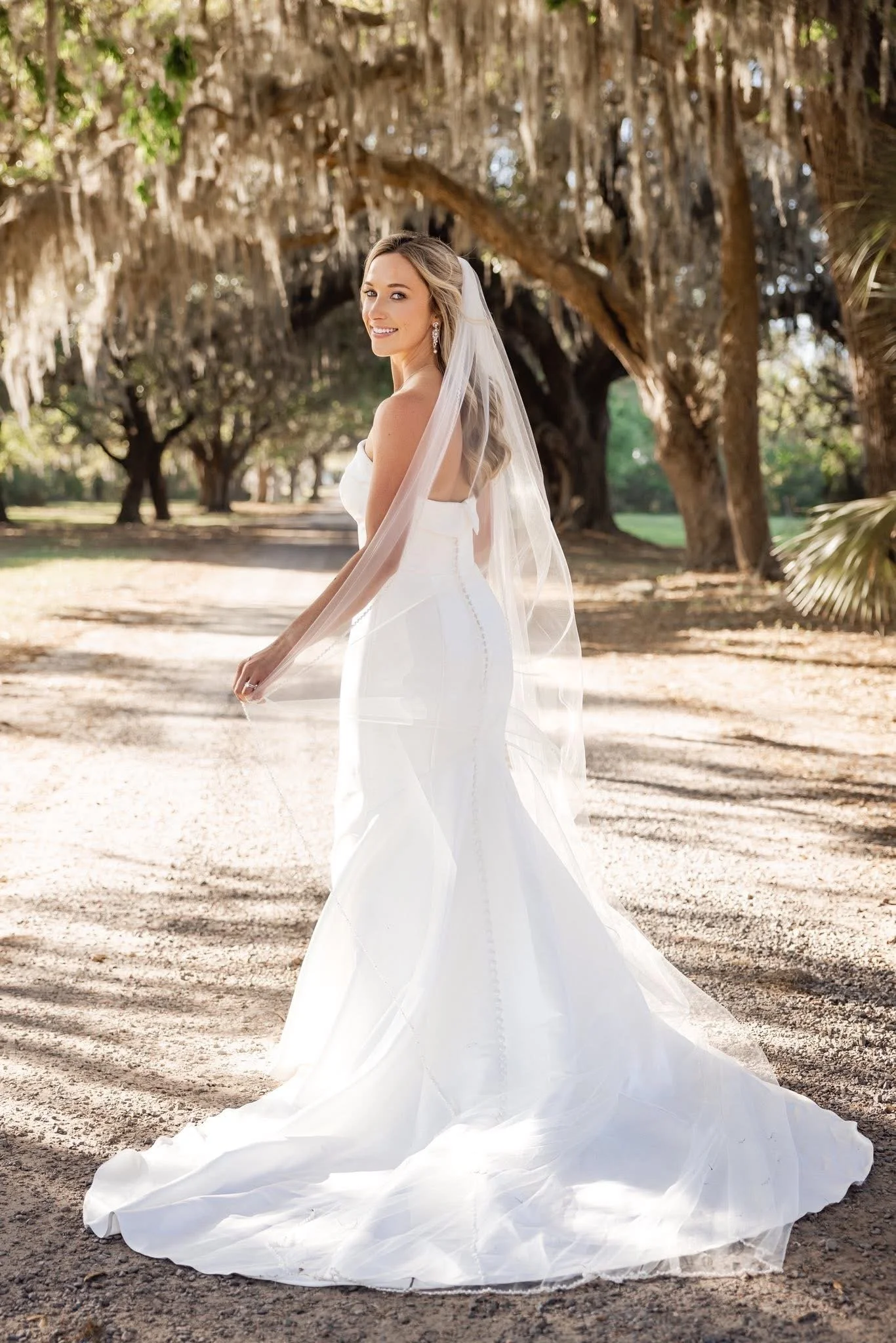 A bride in a white wedding dress and veil standing outdoors on a dirt path under large trees with hanging moss, smiling at the camera.