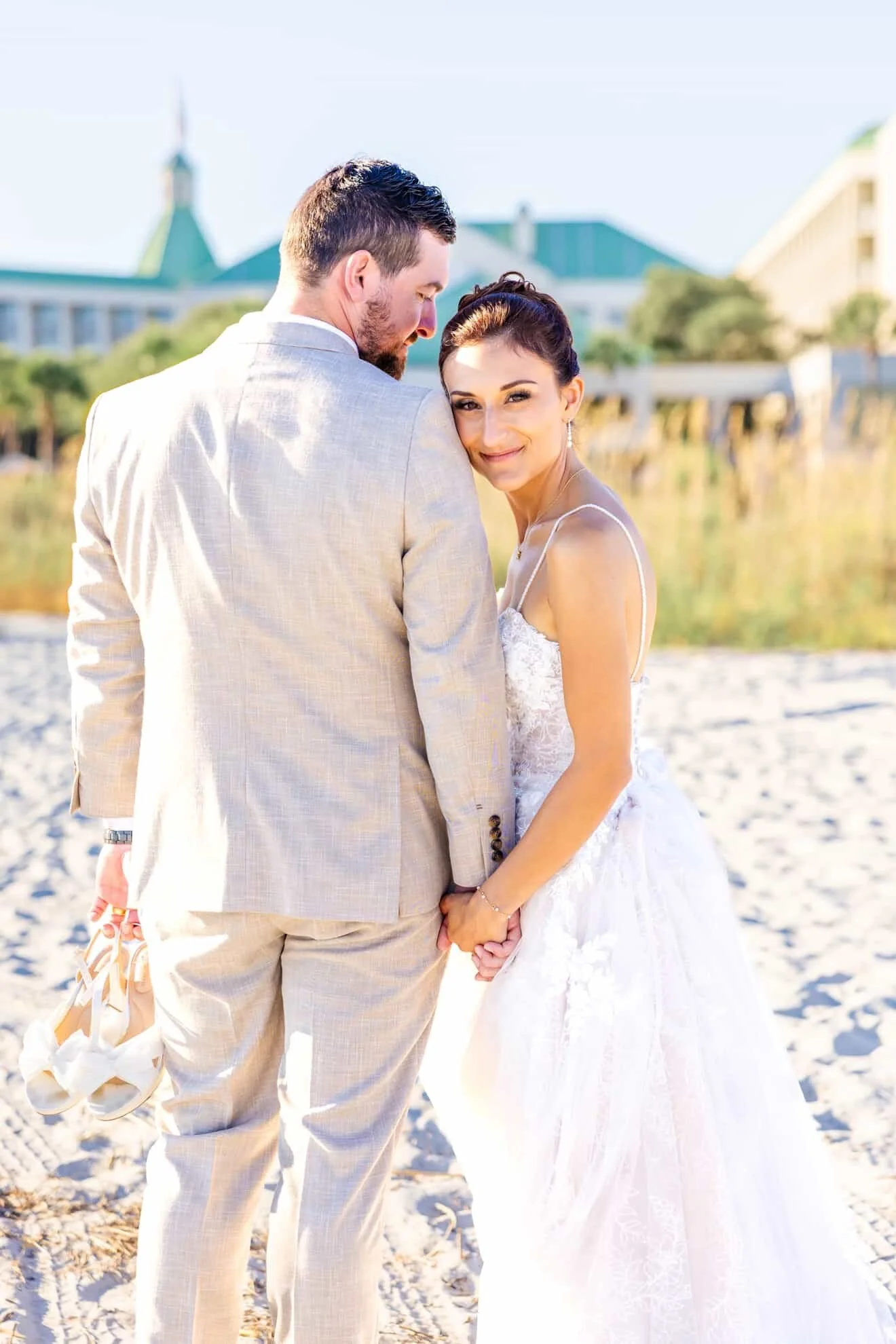 A bride and groom holding hands on the beach during their wedding, smiling with buildings and water in the background on a sunny day.